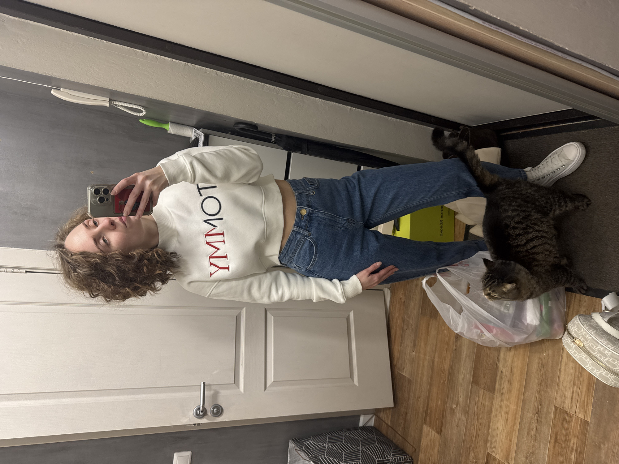 A woman with curly hair is taking a selfie in what appears to be a hallway. She's wearing a white sweatshirt with "YMMOT" printed vertically in red letters and blue jeans. A tabby cat is lying on a plastic bag next to her legs. Other objects visible include a white sneaker, a patterned tote bag, a broom, a black umbrella, and a white telephone on the wall. The location is likely indoors, possibly a home or apartment. No evidence indicates that it is in Perm, Russia.