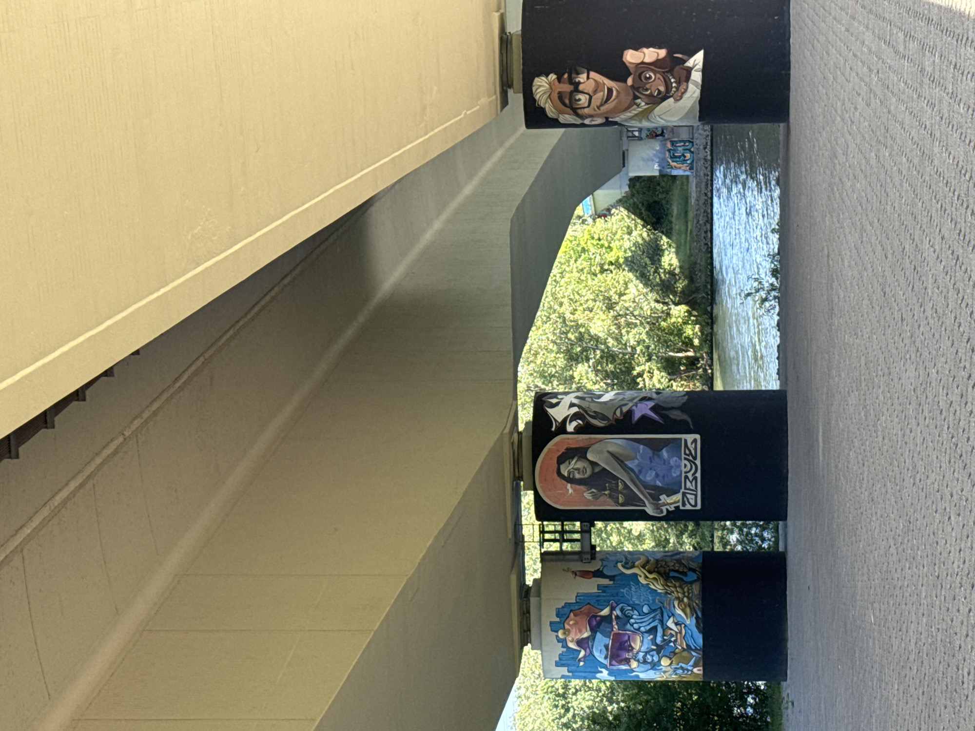 The image displays a detailed view from underneath a concrete bridge in Flörsheim, Germany, showcasing three painted cylindrical bridge pillars. The overhead structure of the bridge consists of light beige-grey concrete beams with visible joints and textures. The ground directly beneath the bridge is a light grey, textured paved surface.
From left to right, the three visible pillars are adorned with distinct murals on their upper sections, with their lower sections uniformly painted black.
1. **Left Pillar:** Features a vibrant, multi-colored graffiti-style mural depicting a blue, skeletal or abstract figure wearing purple sunglasses and a red hat, surrounded by dynamic patterns and a yellowish creature resembling a fish or dragon.
2. **Middle Pillar:** Displays a mural of a woman with long dark hair, blindfolded, holding a set of scales in her left hand and a sword-like object in her right. She is wearing a blue garment. An orange, circular halo-like shape is behind her head, and stylized symbols are present below the figure. A metal grating structure connects the top of this pillar to the left pillar.
3. **Right Pillar:** Presents a cartoon-style mural of a smiling man with white hair, glasses, and a light-colored shirt, holding a small, happy brown dog with a pointed finger.
Behind the pillars, a calm body of water, likely a river, is visible, reflecting the sunlight. The far bank of the water body is densely covered with lush green trees and foliage. In the far distance, beyond the right pillar and across the water, additional concrete structures and some graffiti are visible on what appears to be another part of the bridge or an embankment. The scene is illuminated by natural daylight.