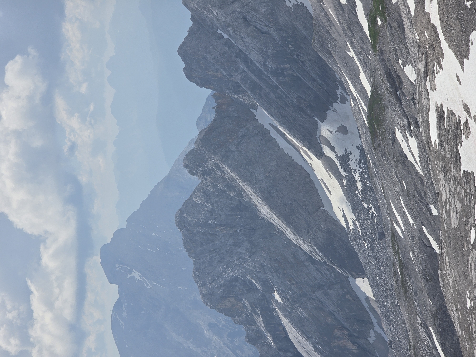 This is a vertical, eye-level shot of a mountain range in Garmisch-Partenkirchen, Germany. The scene is dominated by jagged, gray rock faces, some of which are partially covered in patches of white snow. The mountains recede into the distance, becoming hazy and blue due to atmospheric perspective. In the foreground, the texture of the rocks is rough and stratified. Streaks of snow run down the slopes, contrasting with the dark gray stone.
The sky is a pale blue, with fluffy white clouds scattered across it. The lighting suggests it is daytime, likely late morning or afternoon, with the sun illuminating the scene from an unseen angle. The overall impression is one of a vast, rugged, and natural landscape, with a sense of solitude and the immensity of nature. There are no people or man-made objects visible in the image, emphasizing the wildness of the location. The weather appears clear, with good visibility despite the atmospheric haze in the distance.