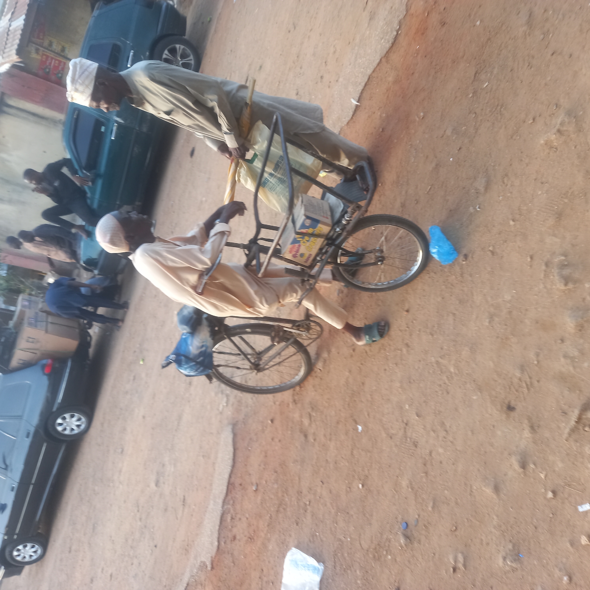 In Dadi Riba, Nigeria, two men are present near a bicycle cart filled with goods. One man, dressed in a light-colored traditional garment and a white cap, sits on the bicycle cart. The other man, also wearing a white cap and a light-colored garment, stands beside the cart, holding a yellow object. The ground is dirt. Several cars are parked nearby. People walk in the background.