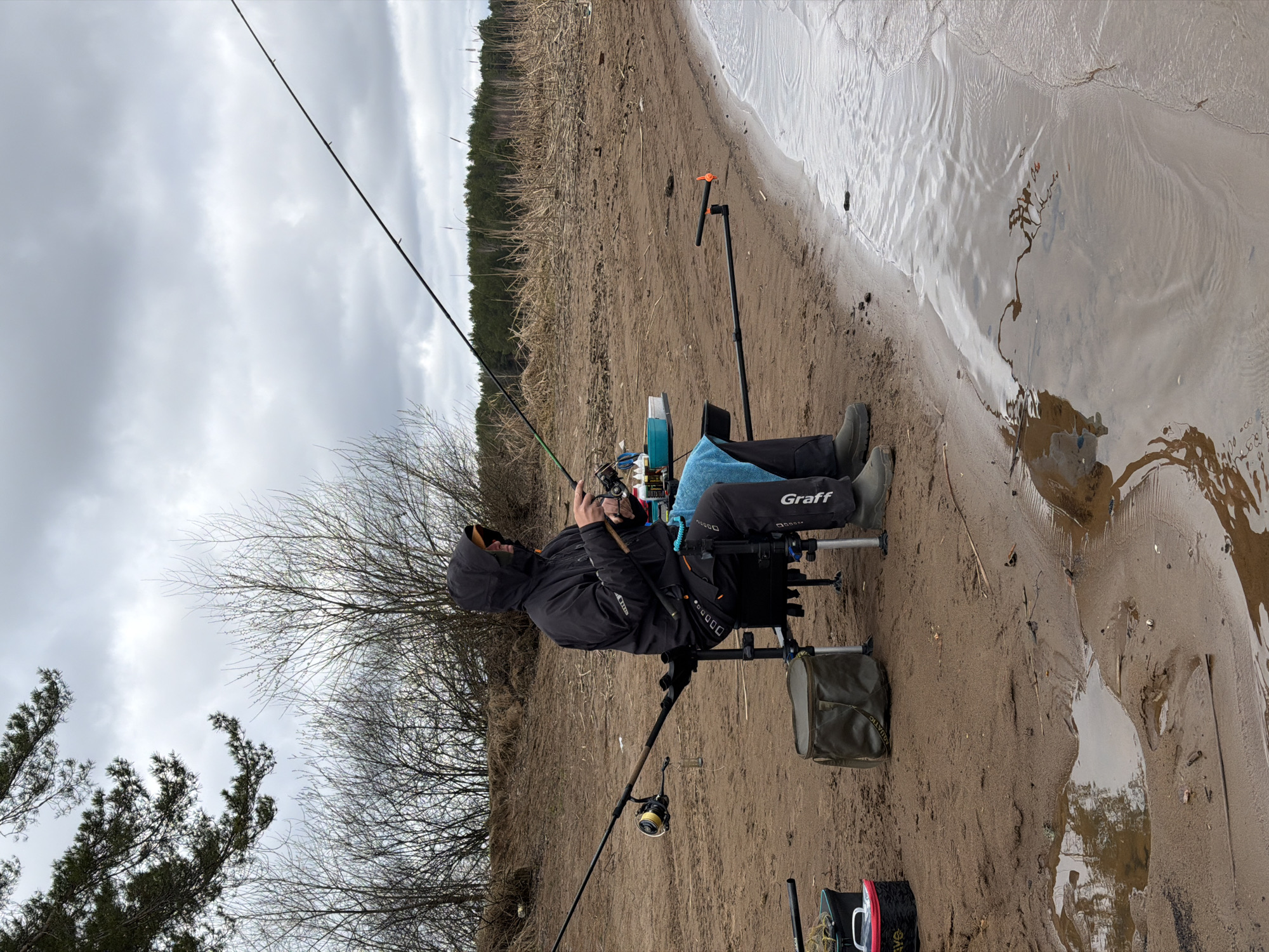 A person is fishing on the shore of a body of water. The person is sitting in a fishing chair, wearing a black jacket with the word "Graff" on the leg, and holding a fishing rod. Two fishing rods are visible. The location is Chaykovsky, Russia. The environment includes water, sand, trees, and a cloudy sky.