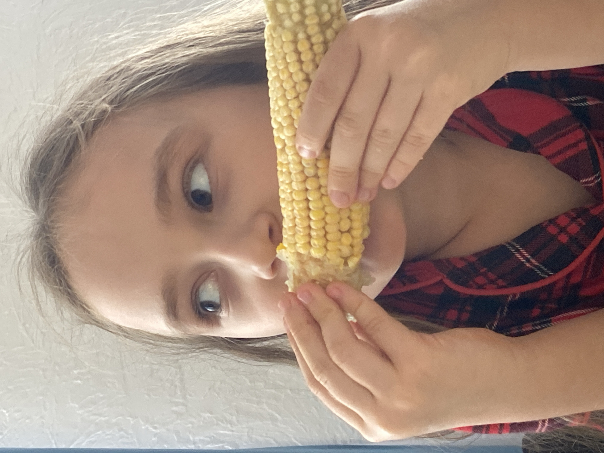 A young girl with light brown hair, wearing a red and black plaid shirt, is eating a cob of corn. She is holding the cob with both hands and looking to her left. The background is a textured white wall. The image appears to have been taken indoors, possibly in a residential setting. No specific location in Oktyabrsky, Russia, can be determined from the image alone.