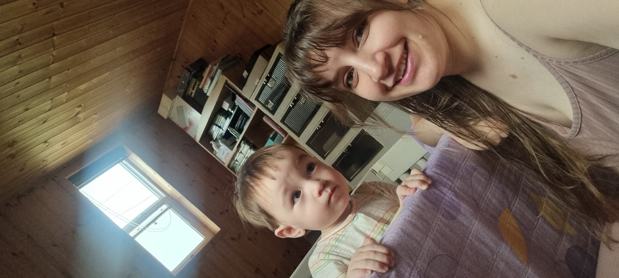 A woman with long brown hair and bangs smiles at the camera while holding a young child, likely a baby, in front of her. The child, with light skin and short brown hair, looks at the camera with a somewhat serious expression, grasping the purple patterned fabric. The background consists of a room with wooden walls and ceiling, suggesting a cabin-like or attic space, and a window on the left side provides natural light. A bookshelf is visible behind them with various items on display. Based on the wooden wall, the house could be located in a rural area. Since there are no distinctive elements to connect the scene to the specific location of Dunnae-myeon, South Korea, the location cannot be determined.