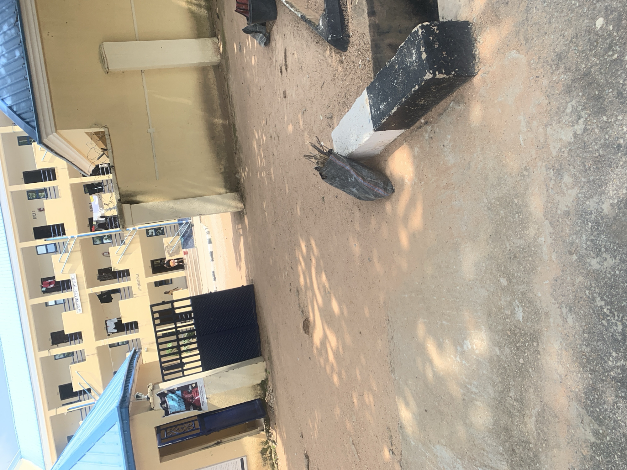 The image depicts a daytime outdoor scene in Jalingo, Nigeria, likely a campus environment. In the background, a cream-colored, multi-story building with a blue roof is visible. The building has multiple windows and balconies, with some laundry hanging out to dry. A black metal gate is in the foreground, partially obscuring the entrance to the building. A poster featuring a person is affixed to the left side of the gate. The ground is a mixture of sand and concrete. In the middle ground, a black trash bag filled with sticks and branches rests against a black and white painted concrete barrier. To the right, a black dustpan and broom are visible, as is a black trash can with red contents. The overall impression is one of an educational or residential facility with some evidence of ongoing maintenance or cleanup.