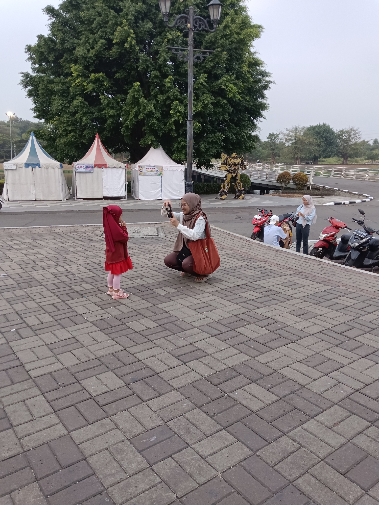 In Jayamukti, Indonesia, a woman is photographing a young girl standing on a paved area. The girl, wearing a red hijab and dress, faces away from the camera. The woman, also in a hijab, kneels and holds a phone to take a picture. Behind them, several tents are set up. In the background, a large tree and a metal transformer statue are visible, along with a bridge and motorcycles.