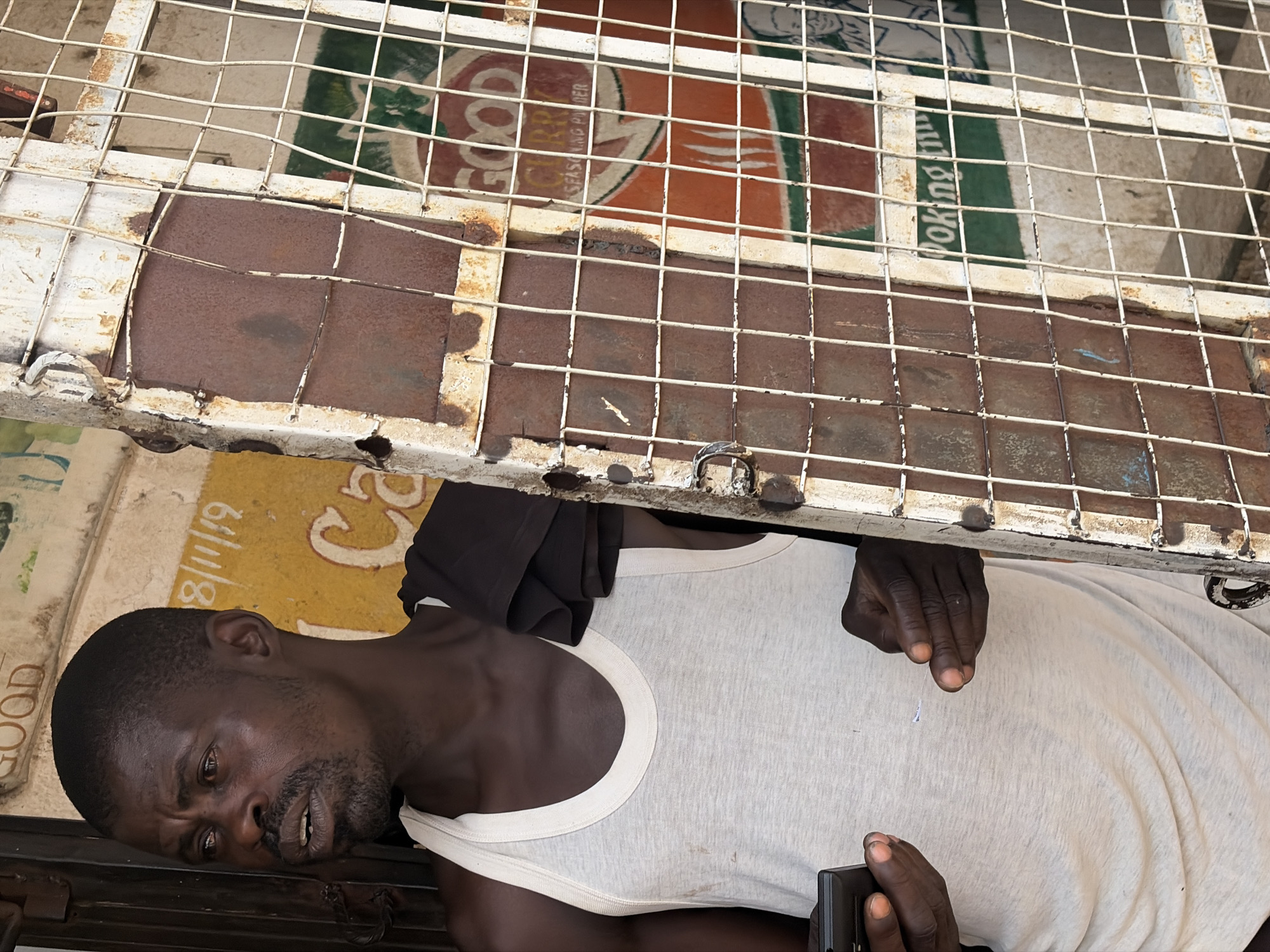 An African man with dark skin, a short haircut, and a goatee is standing in a doorway, looking to his left. He is wearing a white tank top and a black t-shirt is draped over his left shoulder. His right hand is reaching out, and he is holding a black smartphone in his left hand. Behind him, a yellow wall is visible with some writing in black and red. To the right of the man, there is a rusty metal grate with a white frame. Through the grate, a sign with the word "GOOD" is visible in red and white lettering, and below that, the text "SEASONING POWDER" is also visible. Further to the right, a green sign with white text that appears to say "oking in" can be seen. The man is in Jos, Nigeria.