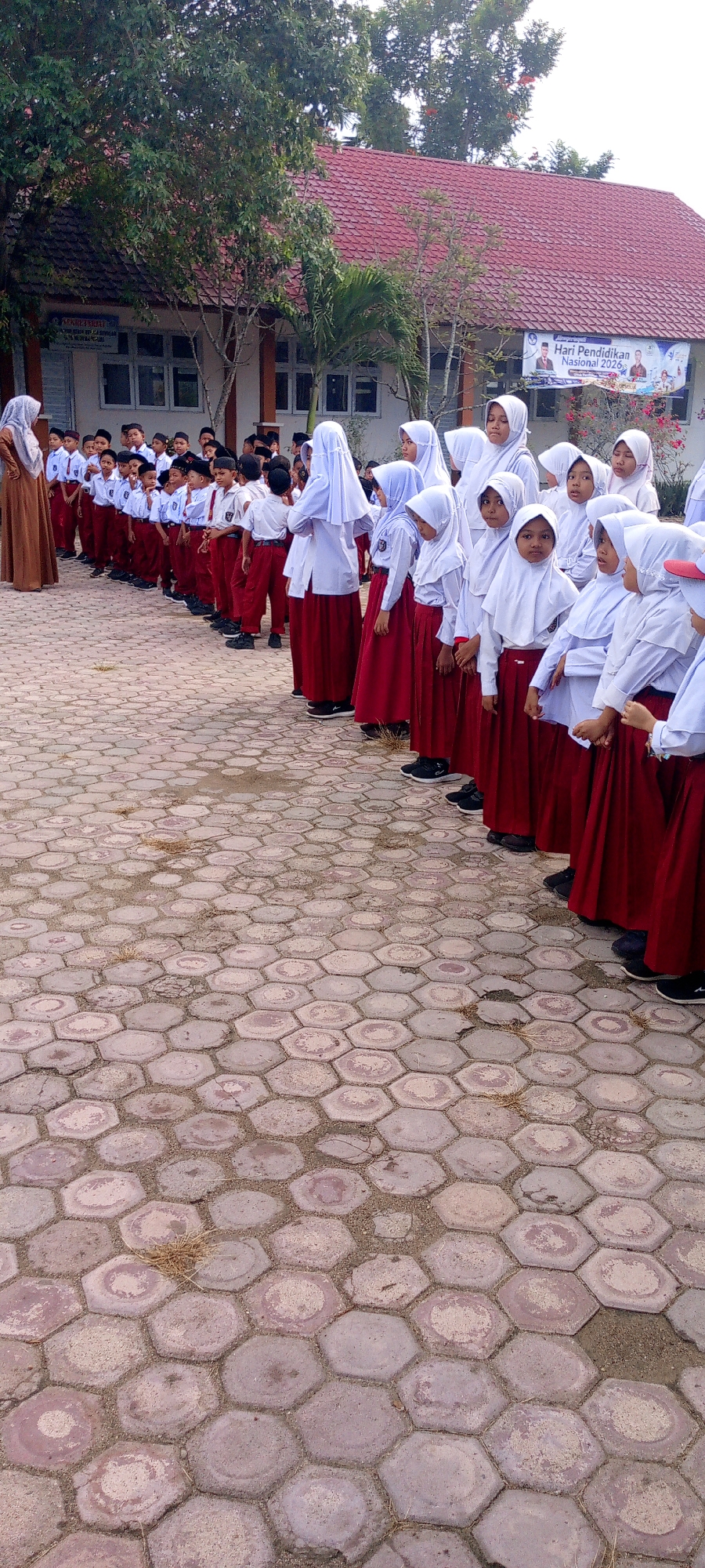 In Baratpayaitek, Indonesia, a group of schoolchildren in uniform are lined up outdoors on a patterned concrete ground. The students are wearing white shirts and red skirts or pants, with some girls wearing white headscarves. They appear to be waiting or listening attentively. Behind them, a building with a red tiled roof is visible, and a banner celebrating "Hari Pendidikan Nasional 2026" (National Education Day 2026) is displayed. Trees and palm trees are scattered around the schoolyard, suggesting an outdoor school setting. The overall atmosphere seems orderly and communal, indicative of a school event or assembly.