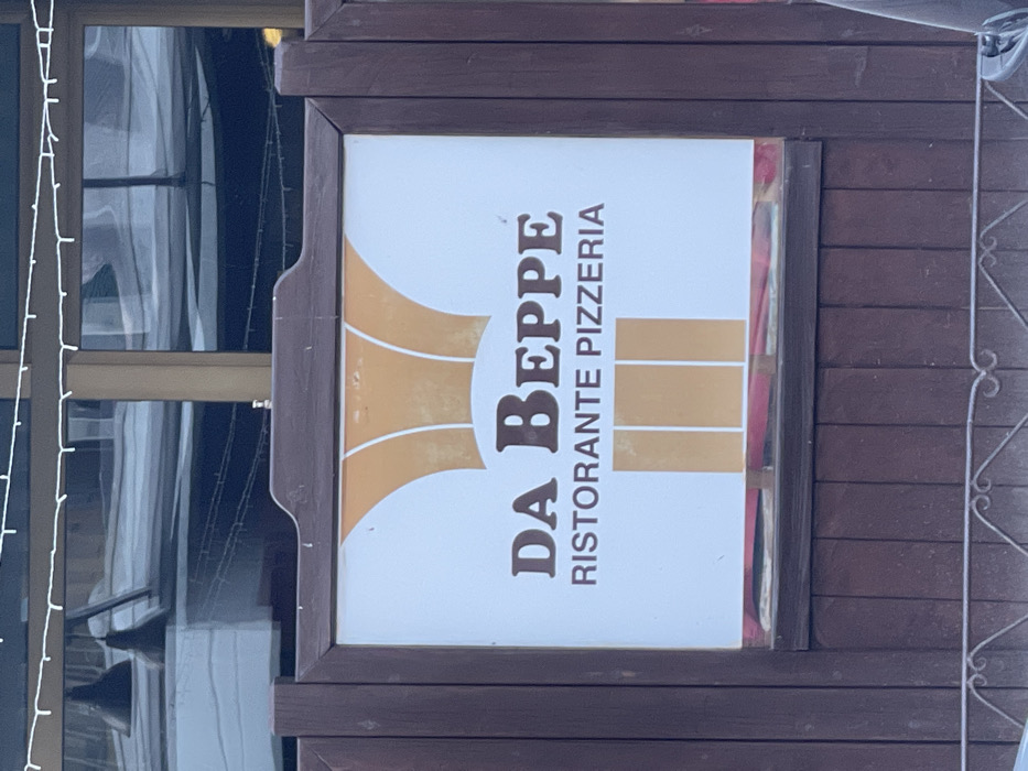 The image displays a sign for an Italian establishment, "DA BEPPE RISTORANTE PIZZERIA," mounted on a dark wooden facade. The sign itself has a white background with a prominent golden or orange abstract design, possibly resembling stylized flames or an abstract chef's hat, with the dark brown text "DA BEPPE" in a larger, bold font, and "RISTORANTE PIZZERIA" in a smaller, sans-serif font below it. The sign is framed by dark brown wood.
To the left of the sign, a large window is visible, reflecting a somewhat blurry scene, possibly showing interior elements or the exterior of an adjacent building. A string of small white lights, likely decorative, hangs vertically along the left edge of the frame, partially obscuring the window. To the right of the sign, a dark, decorative metal railing or gate is partially visible. The overall setting appears to be the exterior of a building, likely a restaurant or pizzeria, consistent with the given location context of Sondrio, Italy. The lighting suggests daytime, possibly overcast, as there are no strong shadows. No people or specific activities are depicted.