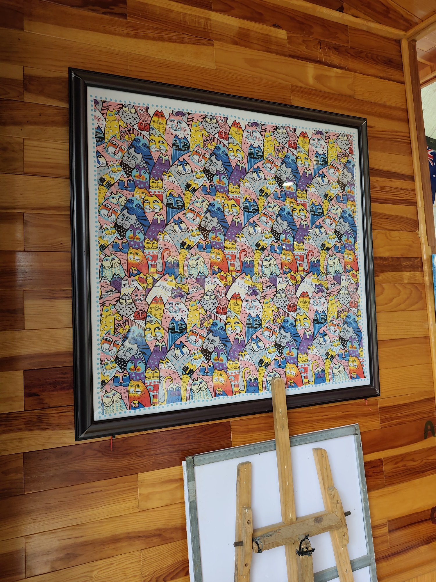 A framed, vibrant artwork featuring a dense pattern of numerous stylized cats is displayed on a wooden paneled wall. The cats are depicted in various colors, including pink, yellow, blue, purple, and orange, with distinct facial features and patterns. A light blue border with small dots frames the artwork. In the foreground, a wooden easel supports a white board. The wooden wall has horizontal planks with a visible grain. To the right of the artwork, a portion of an Australian flag is visible.