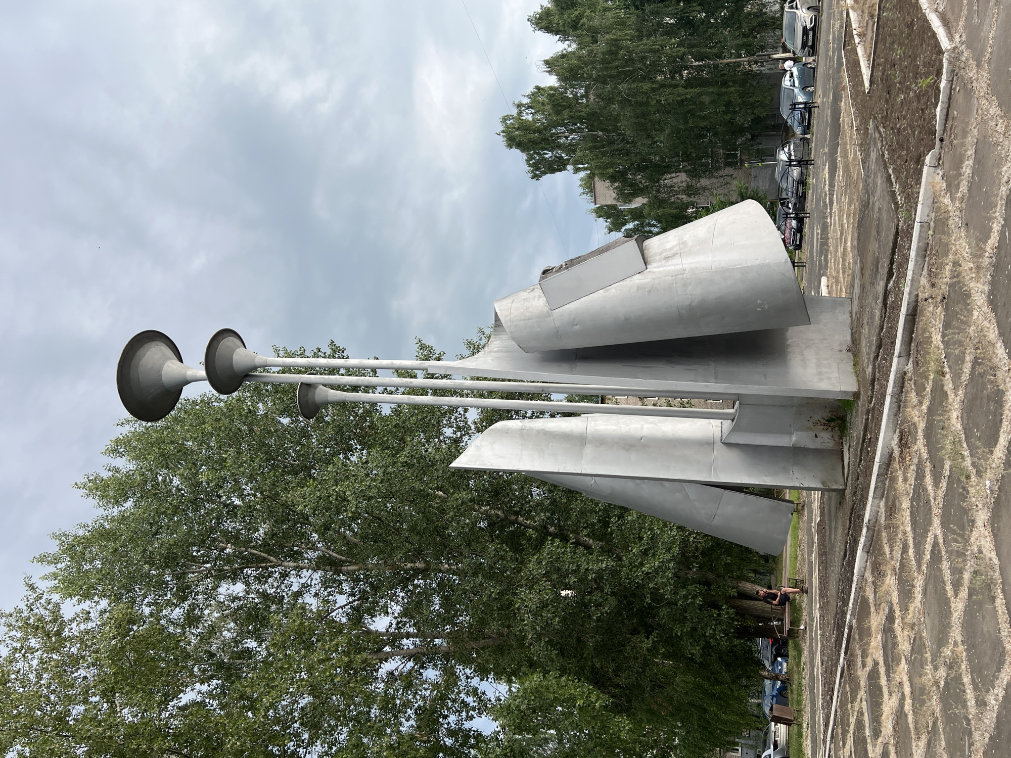 A tall, abstract metal sculpture stands in an open, paved area in Chaikovsky, Russia. The sculpture is comprised of several sharp, angled metallic planes and three slender poles topped with flared trumpet-like shapes. Surrounding the sculpture are mature trees with green foliage. In the background, buildings and parked cars are visible. A person is seated on a bench to the left of the sculpture, looking down. The sky is overcast. The ground in front of the sculpture is paved with square tiles, some of which are cracked and overgrown with dry grass.