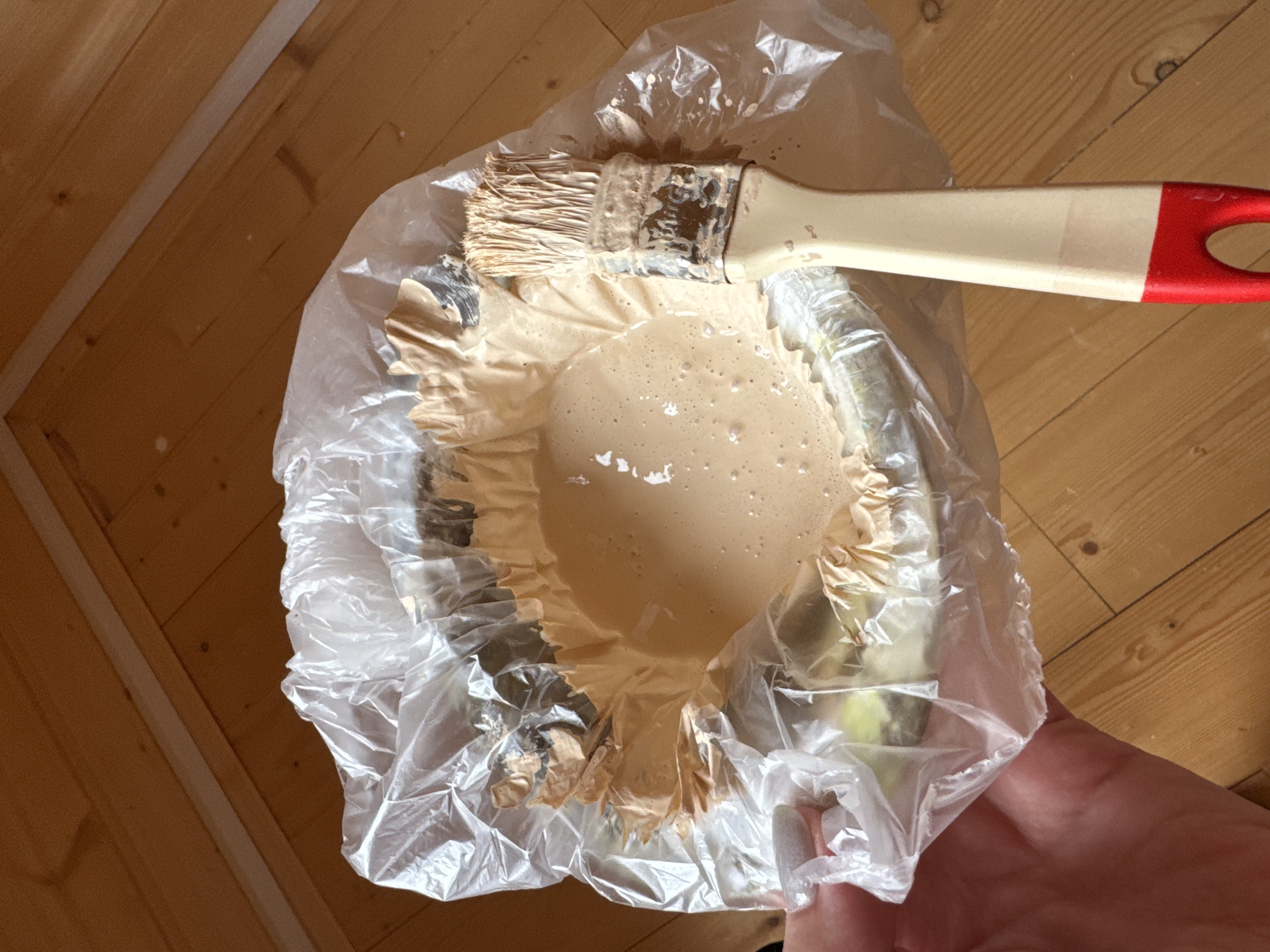 The image shows an indoor scene in Razdolie, Russia, depicting a preparation for painting. A person's hand is visible on the right, holding a clear plastic liner that is used to line a container, likely a paint tray or bowl. The liner is filled with a light, creamy brown paint, which has some small bubbles on its surface. A paintbrush, featuring a creamy handle with a red tip and light-colored bristles, is resting within the paint, thoroughly coated in the same hue. The background reveals wooden floorboards with visible grain and knots. A thin white strip runs diagonally across the wooden surface, possibly a joint filler or trim. The lighting suggests it is daytime. No visible text or discernible emotions are present.