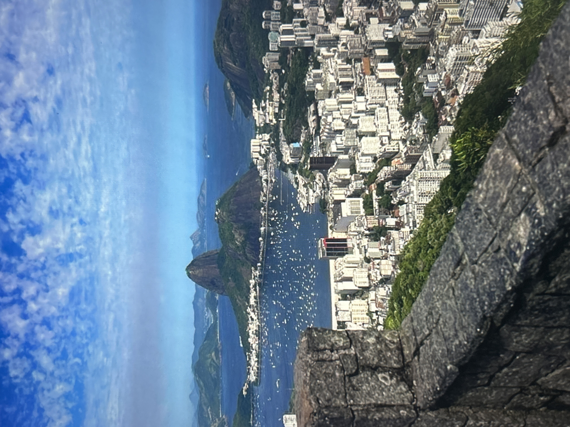The image captures a panoramic view of a coastal city with a prominent mountainous landscape in the background. In the foreground, a stone wall runs along the bottom of the frame, suggesting a viewpoint from an elevated position. Beyond the wall, a dense urban area with numerous buildings stretches out, characterized by a mix of architectural styles and heights, many of which appear to be residential or commercial structures.
The city is situated along a bay or harbor filled with a multitude of boats, indicating a busy maritime activity. The water is a deep blue, contrasting with the lighter tones of the buildings. Dominating the middle ground is a distinctive, rounded mountain covered in lush green vegetation. Further in the distance, a chain of smaller, similarly vegetated mountains extends along the horizon, with a hazy blue sky overhead dotted with scattered white clouds. The overall impression is of a vibrant, densely populated city nestled within a scenic natural environment.