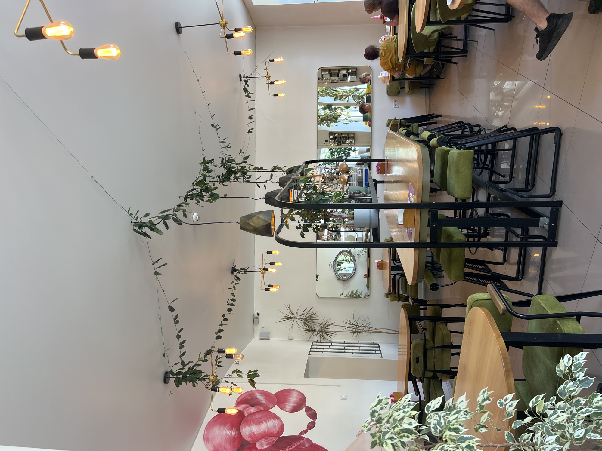 The image depicts the interior of a modern cafe, possibly located in Odesa, Ukraine. The space is brightly lit by natural light and a series of artistic pendant lamps with exposed Edison bulbs. Greenery, including hanging plants and potted plants, is strategically placed throughout the cafe, adding a touch of natural ambiance.
The cafe features several tables, predominantly with light wooden tops and dark metal frames, complemented by plush green upholstered chairs and banquettes. The seating arrangement suggests a capacity for both small and larger groups. In the background, mirrors are mounted on the walls, reflecting the cafe's interior and creating a sense of spaciousness.
Several patrons are visible, engaged in conversations and enjoying their time. On the left side of the frame, a large, abstract mural of red, balloon-like shapes adorns the white wall. The flooring is a polished light-colored tile, reflecting the overhead lighting and furniture. The overall aesthetic is contemporary and inviting, with a focus on comfort and style.