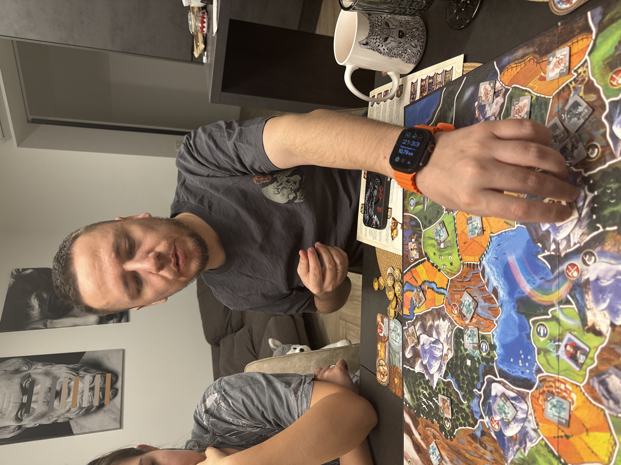 The image captures an indoor scene, likely in a home in Krasnoyarsk, Russia, depicting two individuals engaged in playing a board game during the evening.
The primary subject is an adult man with a beard, wearing a dark grey t-shirt, who is looking towards the viewer and appears to be speaking or explaining something. His right arm, adorned with a smartwatch featuring a vibrant orange band, rests on the table as his hand interacts with the board game. The smartwatch clearly displays the time "21:33" (9:33 PM), a distance of "10,79 KM", and the number "830", possibly a step count or other metric.
Partially visible in the lower left is an adult female, also in a dark grey shirt, leaning on the table and observing the game, with her attention seemingly focused on the board.
The central focus of the activity is a colorful board game spread across a dark table. The game board features a detailed map with various regions and numerous game tokens and cards. Circular tokens resembling coins are visible, alongside square and rectangular character or territory tokens. Some tokens bear text, such as "Атлантида" (Atlantida), "НОАЛ" (NOAL), and "ТРИТОНЫ" (TRITONS), suggesting a game theme involving civilizations or mythological figures.
Other objects on the table include a white mug decorated with an intricate black-and-white animal design (possibly a fox or wolf) and a smartphone case with the word "BERSERK" visible. In the background, a small dog is partially visible, peeking over the back of a sofa or chair. The wall behind the man features two framed black-and-white art pieces, one depicting a classical bust with strips of tape placed over its mouth. The scene conveys a relaxed and engaging atmosphere, characteristic of a casual game night.