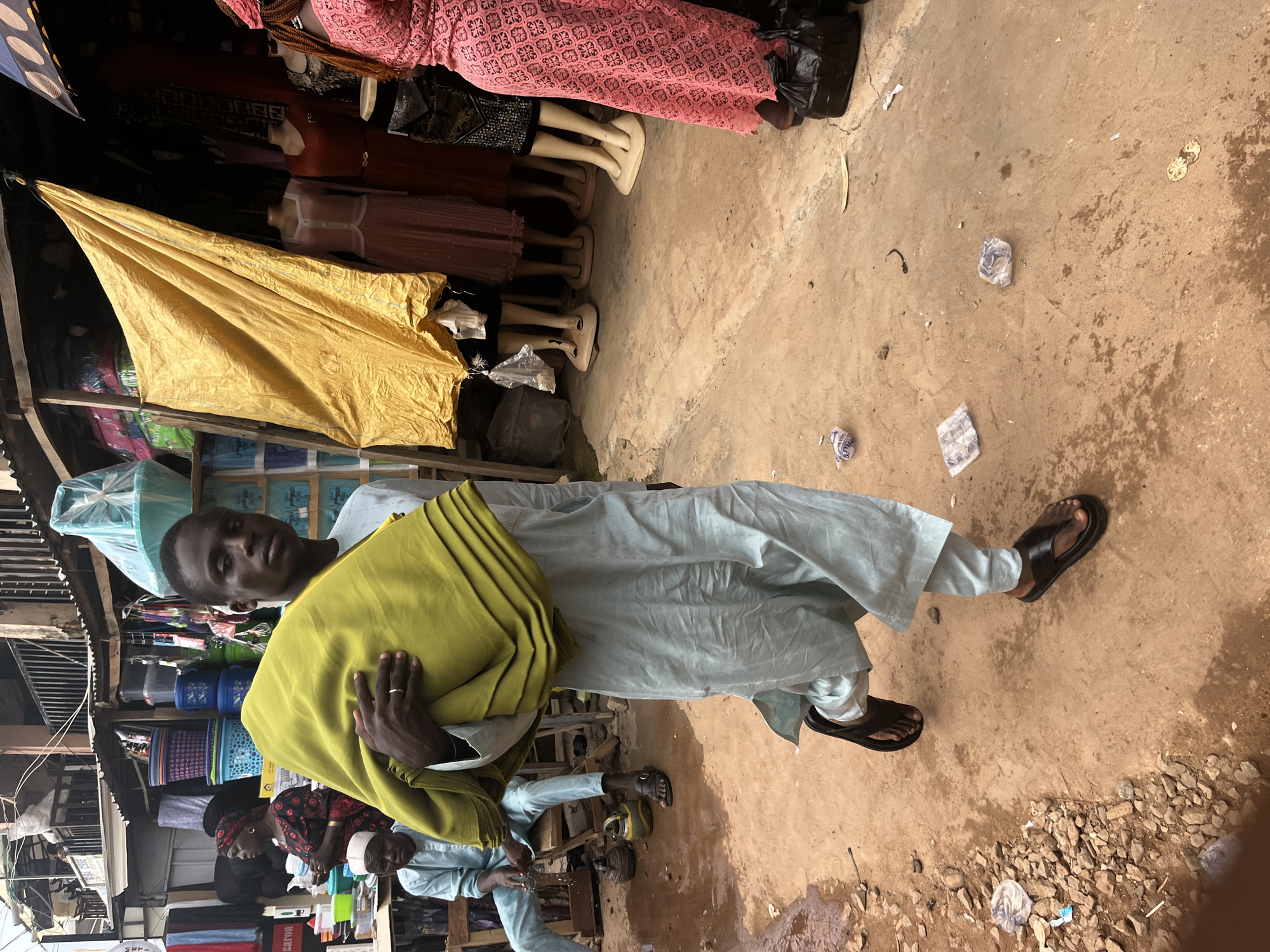 This image depicts a street scene in Jos, Nigeria. The foreground is dominated by a man wearing a light blue traditional outfit and a green headwrap. He is holding a large piece of green fabric. He has dark skin and is looking towards the camera with a neutral expression. The ground is a mix of packed earth and scattered gravel. There are some pieces of plastic litter on the ground, including some wrapped items.
Behind the man, a market stall is visible, displaying various items. Mannequins wearing traditional clothing, including a pink lace dress, are present. A yellow tarp is draped over some of the goods. Further in the background, shelves are stocked with what appear to be containers and textiles. Another person, a woman wearing a dark patterned top and a red headscarf, is visible in the background, as well as a man in a white cap and traditional attire.
The lighting suggests it is daytime, possibly morning or afternoon, with no strong shadows indicating harsh midday sun. The overall scene appears to be a busy, open-air market. The presence of various textiles, clothing on mannequins, and stacked goods points to a commercial environment. The clothing worn by the individuals is indicative of traditional Nigerian attire.