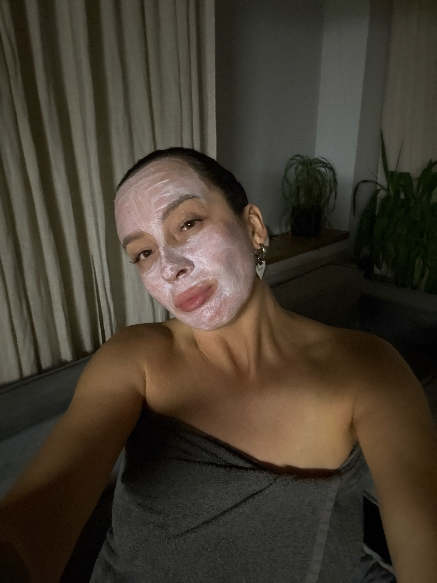 A woman is taking a selfie while wearing a white face mask. She has dark hair pulled back, and is wearing a grey towel wrapped around her. She is wearing a heart-shaped earring. In the background, there are beige curtains and two potted plants. The lighting is dim. The image was taken in Chaikovsky, Russia.