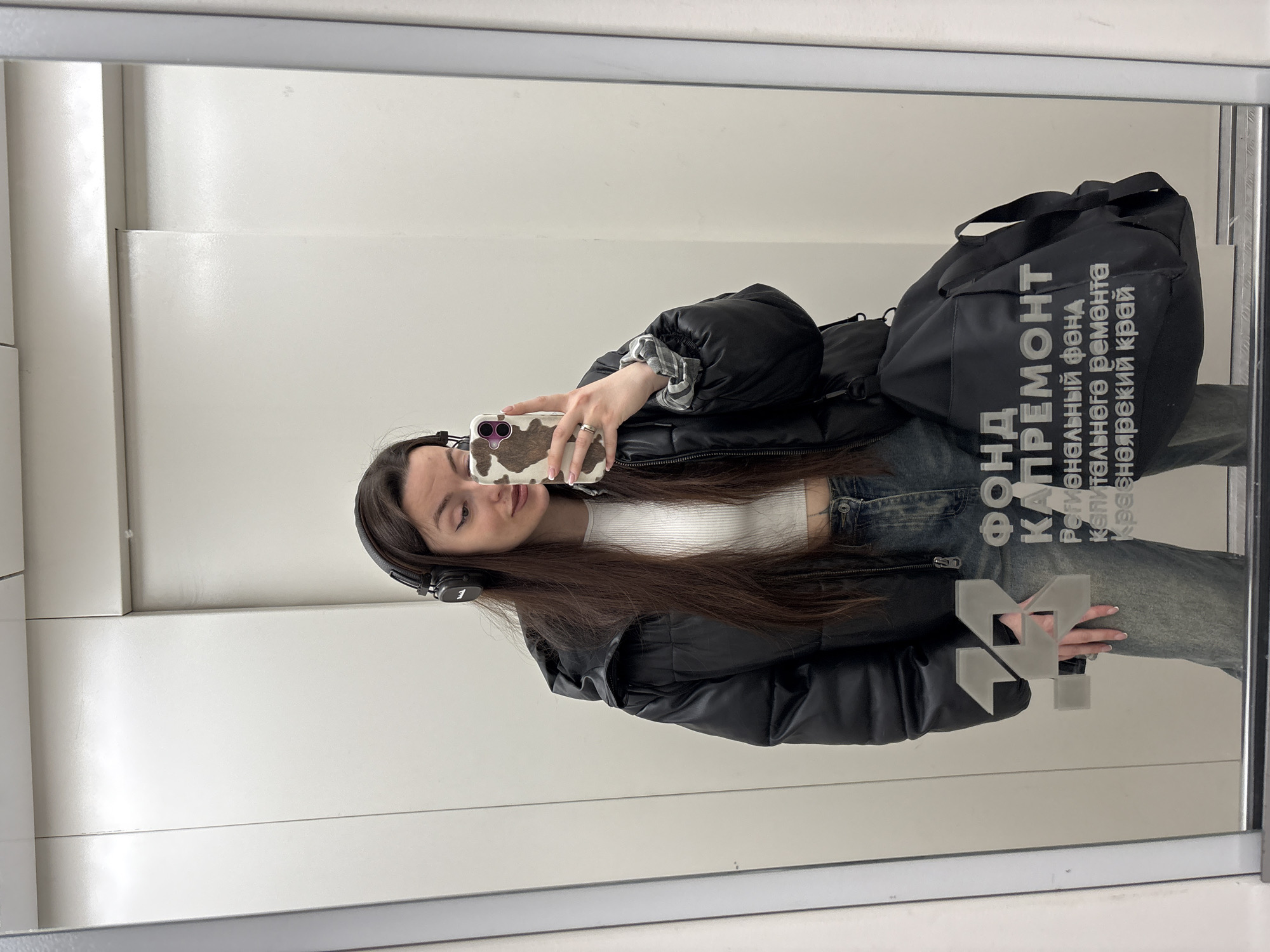 A young woman is taking a selfie in a mirror, likely in an elevator or a similar enclosed space with a bright white interior. She is wearing headphones, a white ribbed top, jeans, and a black puffer jacket. She has a large black bag slung over her shoulder. Her phone has a cow-print case. The mirror reflects signage, reading "Фонд капремонт регионального фонда капитального ремонта Красноярский край," which translates to "Capital Repair Fund of the Regional Capital Repair Fund of the Krasnoyarsk Territory." This indicates the location is Krasnoyarsk, Russia. There is also a "24" logo visible on the mirror, partially obscured by her hand.