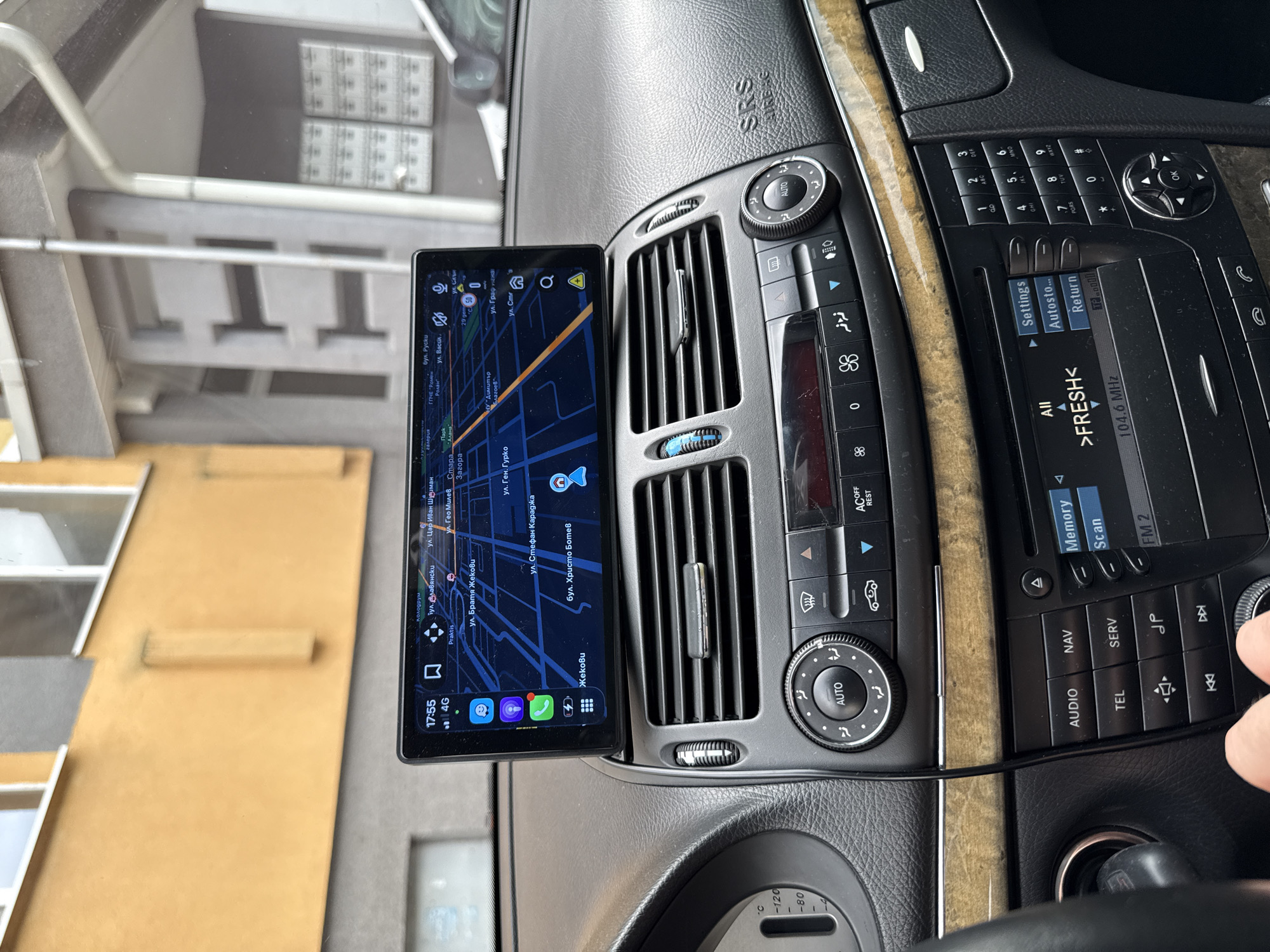 A close-up interior shot focuses on the center console of a car, displaying a tablet attached to the air vents displaying a map. The map indicates the car is in the Kolodrum district of Stara Zagora, Bulgaria, near "Praktis" and streets such as "Bul. Dliavyanski," "Ul. Tsar Ivan Shishman," "Ul. Bratya Zhekovi," and "Ul. Gen. Gurko." The car's radio display shows the frequency "104.6 MHz" and station ">FRESH<." Adjacent buttons include "AUDIO," "NAV," and "TEL." The car’s climate control system and emergency systems are also visible. A hand appears on the right side of the image, possibly interacting with the car's controls. The dashboard is wood and black, with the SRS AIRBAG label visible above the vents.