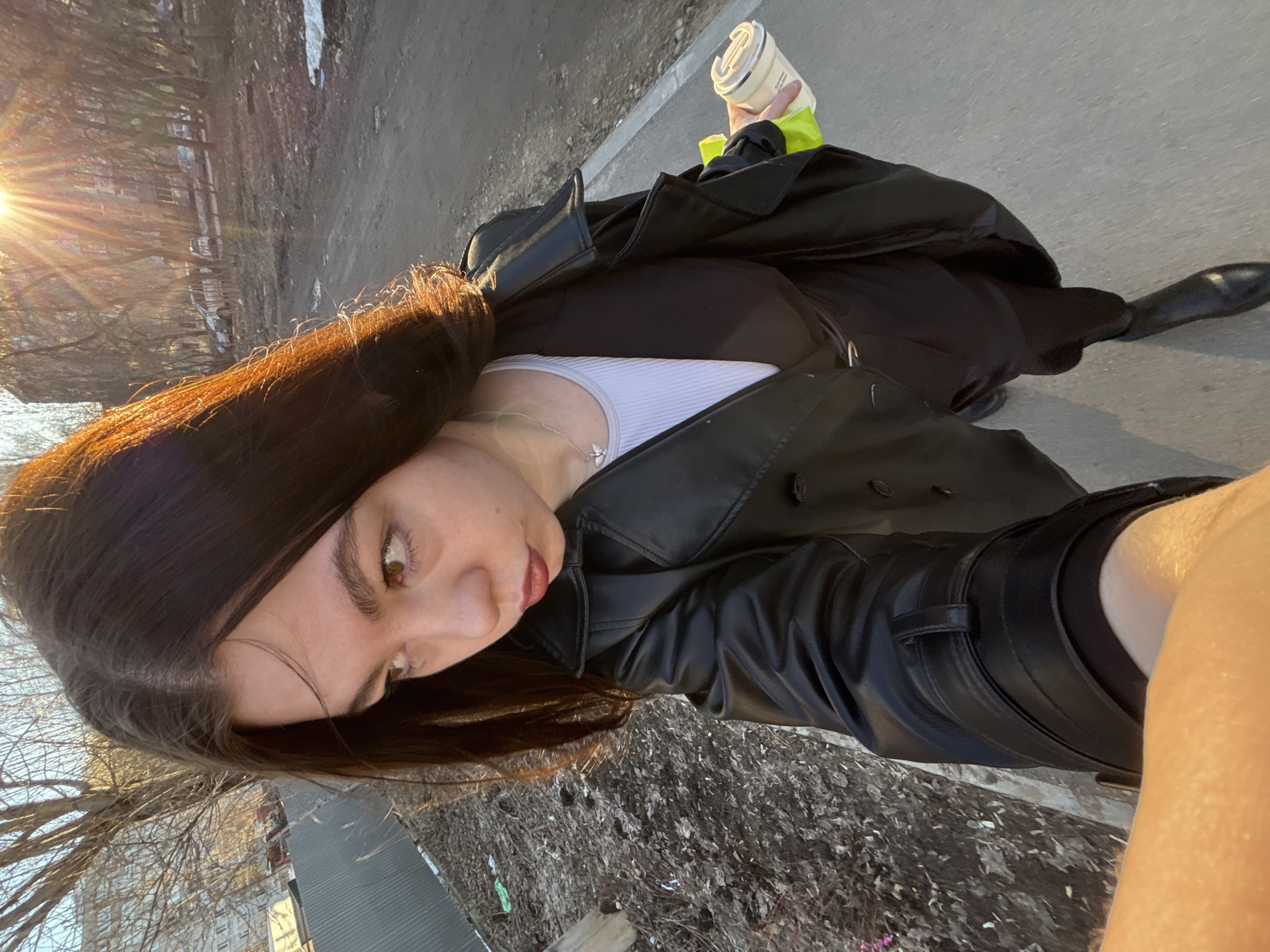 A young woman in a black leather trench coat stands on a sidewalk, holding a beverage in her right hand. She is facing toward the camera with sunlight shining on her dark brown hair. She is wearing a white tank top under a black top, and black pants. A necklace is visible around her neck. The sidewalk is adjacent to a grassy area with a building partially visible in the background. The location is likely Perm, Russia.