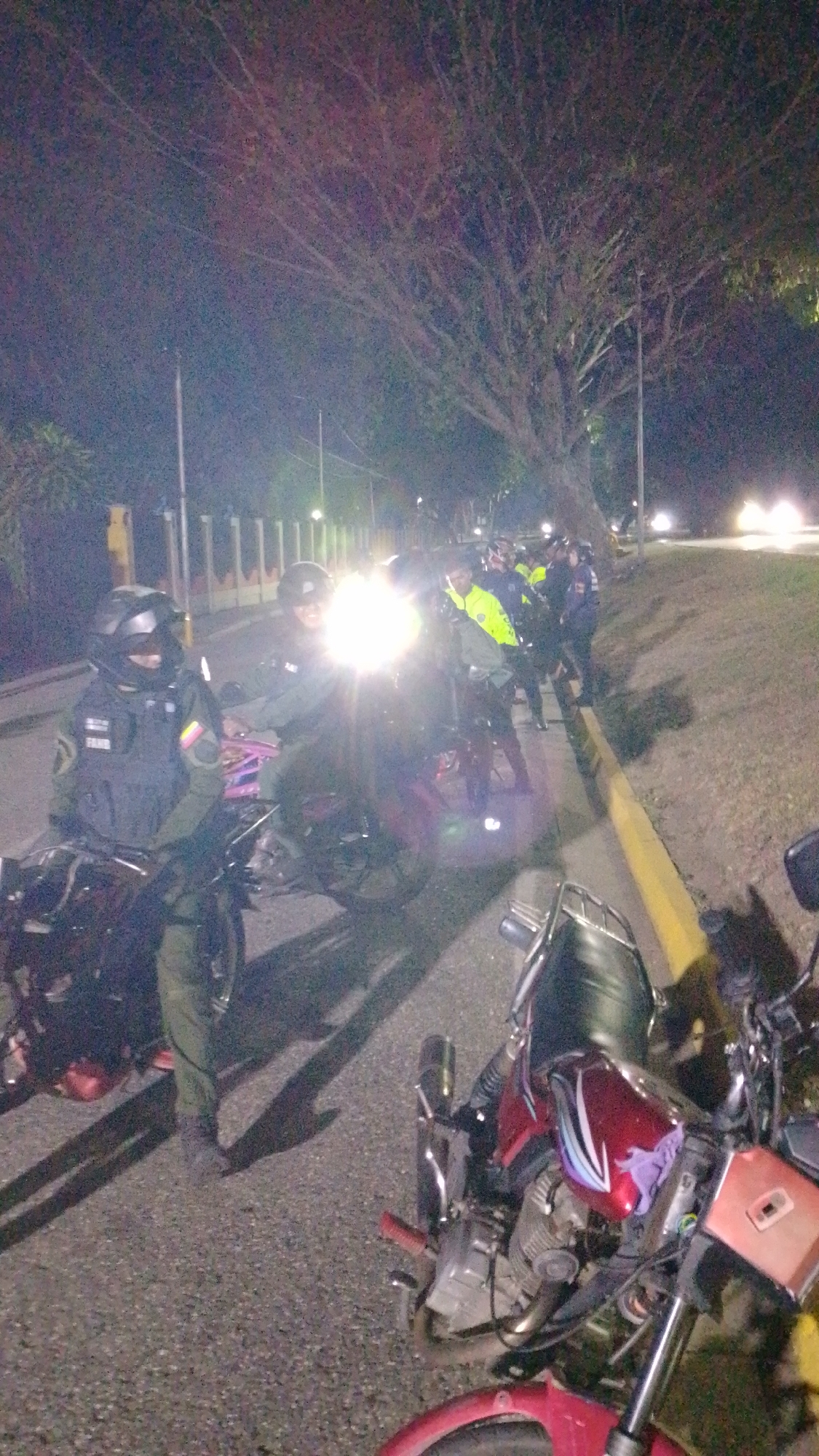 At night in Tinaquillo, Venezuela, a group of uniformed police officers are standing near a row of motorcycles parked on the side of a paved road. The officers are wearing helmets and body armor. Some are standing next to their motorcycles, while others are gathered in a group. One motorcycle has a red gas tank with a design. Streetlights illuminate the scene, casting shadows on the road. A large tree looms over the scene. Other vehicles are visible in the distance.
