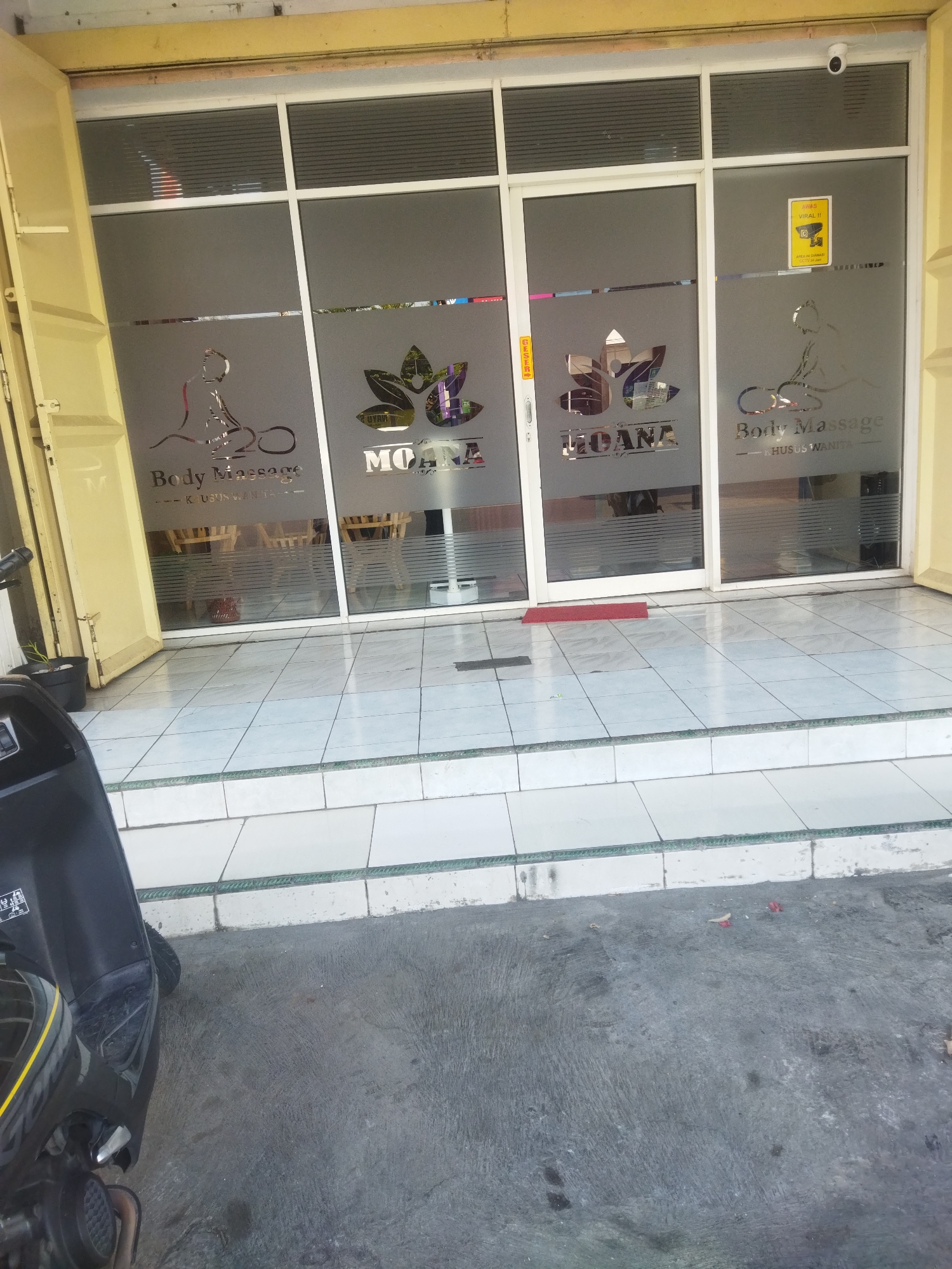 The image shows the exterior of a business establishment, likely a spa or massage parlor, in Sukoharjo, Indonesia. The storefront features a double glass door entrance with frosted glass decals. The decals include stylized human figures, lotus flower motifs, and text that appears to read "MOANA" and "Body Massage - KHUSUS WANITA" (which translates to "For Women Only"). A yellow security camera is visible mounted on the top right corner of the door frame. A red doormat is placed on the tiled floor in front of the entrance. To the left of the entrance, a black scooter is parked partially in the frame. The building's facade is painted in yellow. The ground in front of the establishment is paved with gray concrete. The overall environment suggests a commercial area in an Indonesian city.
