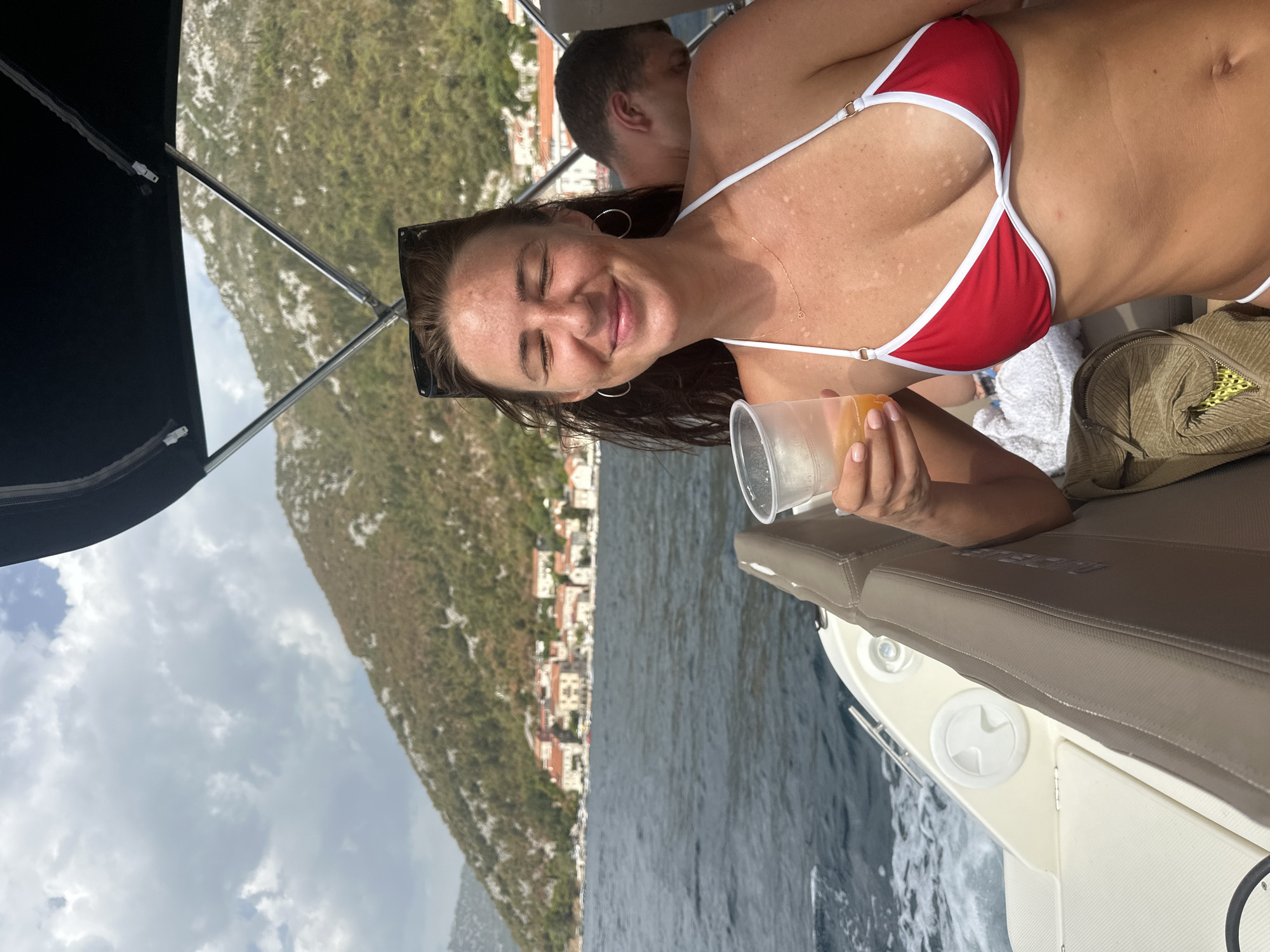 A woman in a red bikini holds a drink while on a boat in Perast, Montenegro. The woman has dark hair and is wearing large hoop earrings and a delicate necklace. She has her eyes closed and is smiling. In the background, a man is visible, leaning back on the boat. The boat is on the water, with a coastal town and hills in the distance. The sky is cloudy.