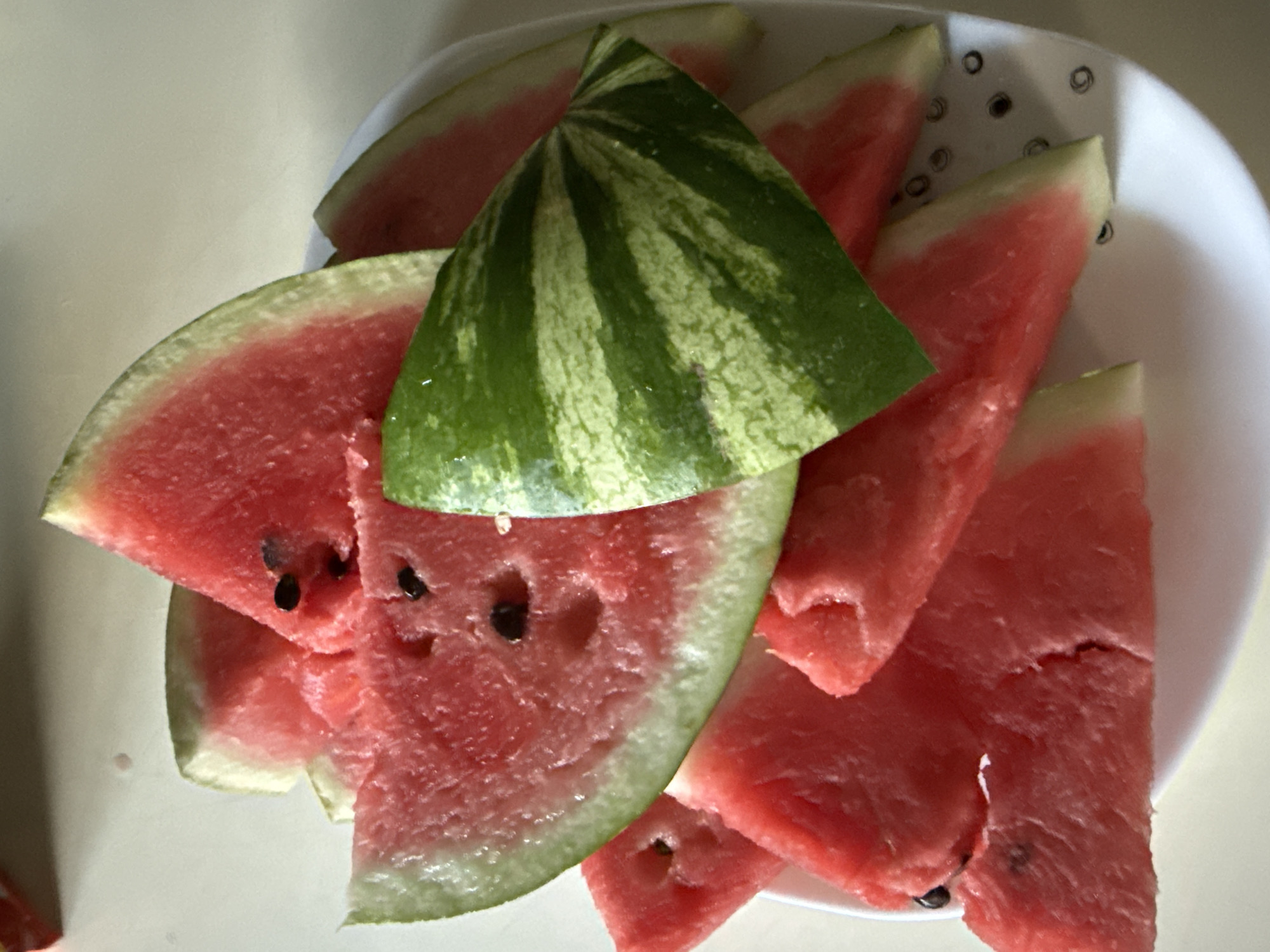 A close-up, overhead shot showcases a white plate piled high with sliced watermelon. The watermelon slices are a vibrant pink with some exhibiting black seeds. One particularly large slice is angled prominently, revealing its striped green rind. The plate itself has a faint pattern of small, dark circles. The background is a light-colored, out-of-focus surface. The scene does not provide any information about the city of Okhtyrka, Ukraine.