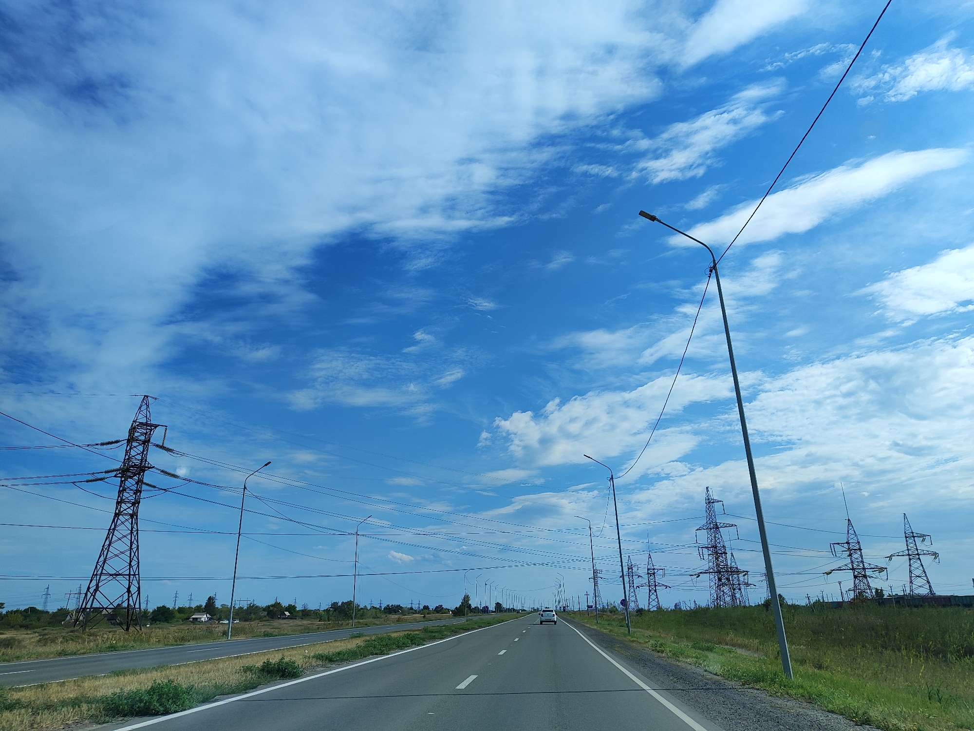 This is a photograph taken from a moving vehicle on a highway near Aksu, Kazakhstan. The sky is a vibrant blue, dotted with scattered white clouds, some of which are wispy and stretched across the expanse. The highway stretches into the distance, with a white line marking the lanes. On either side of the road, there are grassy fields and trees. Numerous tall, metal power line towers, some of them very large and lattice-like, are visible in the landscape, along with street lights lining the highway. A white car is driving ahead on the highway. The overall scene depicts a modern, somewhat industrial, roadside environment under a bright, cloudy sky.