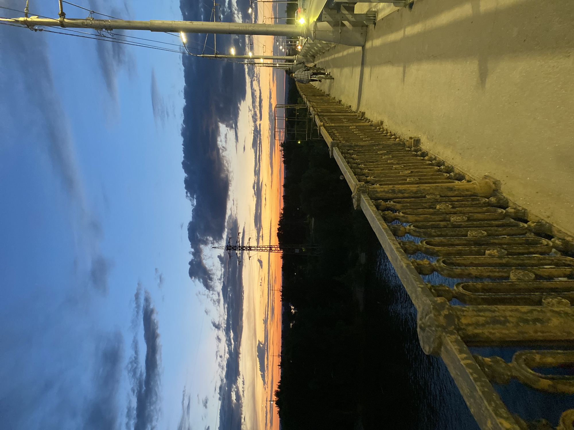 The image captures a twilight scene from a bridge in Novokuybyshevsk, Russia, overlooking a body of water and a distant treeline. The sky displays a gradient of blues and oranges, with scattered dark clouds and the last vestiges of sunset. A person on a bicycle is visible on the bridge, followed by another cyclist. The bridge has a concrete walkway and a decorative, weathered metal railing. Streetlights illuminate the path, casting long shadows. In the background, power lines and transmission towers are silhouetted against the colorful sky, and the dark shapes of trees line the far bank of the water.