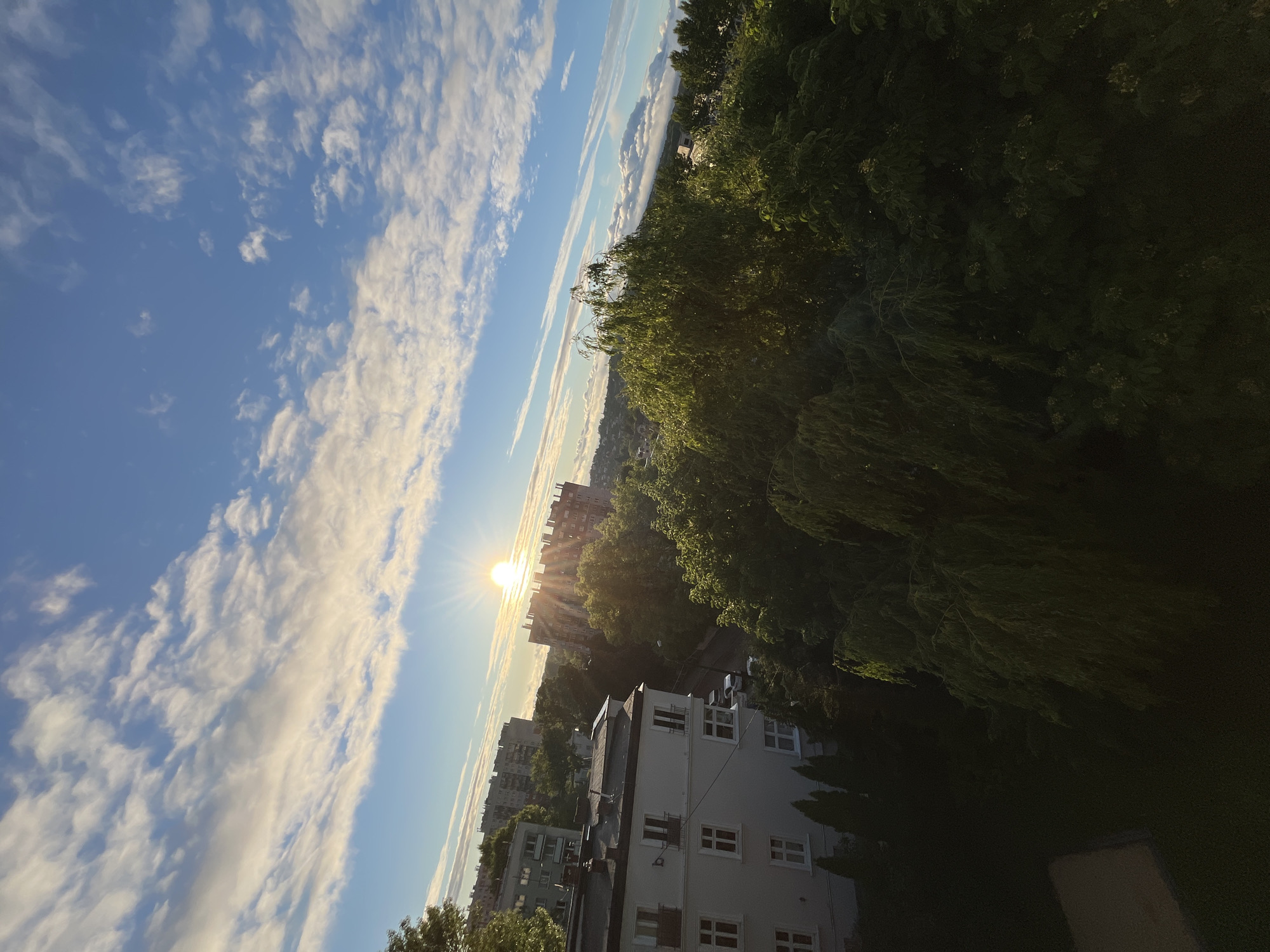 An elevated, outward-looking photograph captures a vibrant sunrise or sunset over a residential area. The sky occupies the upper two-thirds, featuring a deep blue hue with scattered white, wispy clouds. A prominent, intensely bright sun, exhibiting a starburst effect, is positioned in the upper-middle left, casting long rays and warm light across the scene.
In the foreground, a significant portion of a light-colored multi-story building with multiple windows and balconies is visible on the left. The windows are rectangular with dark frames. Below this building, a light-colored rectangular surface is partially visible at the bottom left. The middle ground is dominated by a dense canopy of dark green trees, with some upper leaves catching the direct sunlight. A street or road with several parked white vehicles is discernible between the trees and buildings.
Further into the mid-ground and background, various multi-story apartment blocks, some with visible windows and balconies, are scattered amidst more greenery. These buildings appear to be of a distinct architectural style from the foreground structure. The distant horizon reveals a sprawling urban landscape under a light haze or lower cloud layer. No individuals are present in the image. The scene is illuminated by the low-angle sunlight, creating a dynamic interplay of light and shadow, particularly on the foliage and building facades. The overall location is an urban setting within Unknown, Unknown.