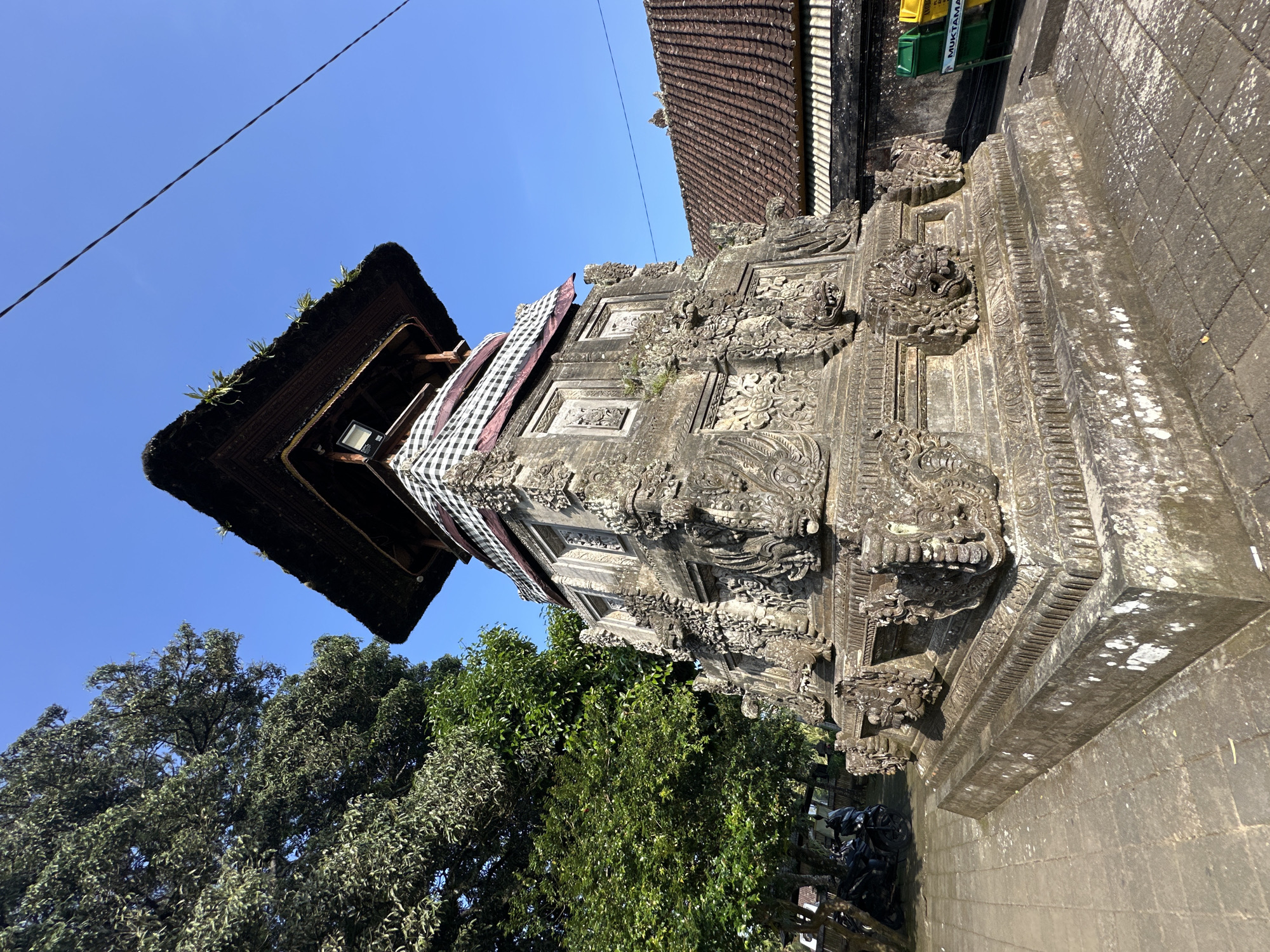 The image shows a detailed, multi-tiered stone structure with intricate carvings, topped with a thatched roof. The structure appears to be a temple or monument, possibly located in a tropical environment given the presence of lush green trees and a clear blue sky. The stone is weathered, showing patches of moss and lichen, indicating age and exposure to the elements. The lower section of the structure is a substantial stone base with decorative elements, possibly depicting mythological creatures. A black and white checked cloth is draped around the upper part of the stone section, just below the thatched roof. The roof itself is dark, possibly made of straw or reeds, and has some vegetation growing on it. In the background, there is a tiled roof of a building, suggesting an inhabited area. To the left of the structure, there are motorcycles parked, and to the right, two recycling bins, one green and one yellow, are visible. The ground is paved with stone tiles. The scene is well-lit by natural sunlight, casting shadows on the structure and pavement. The location is identified as Bangli, Indonesia.