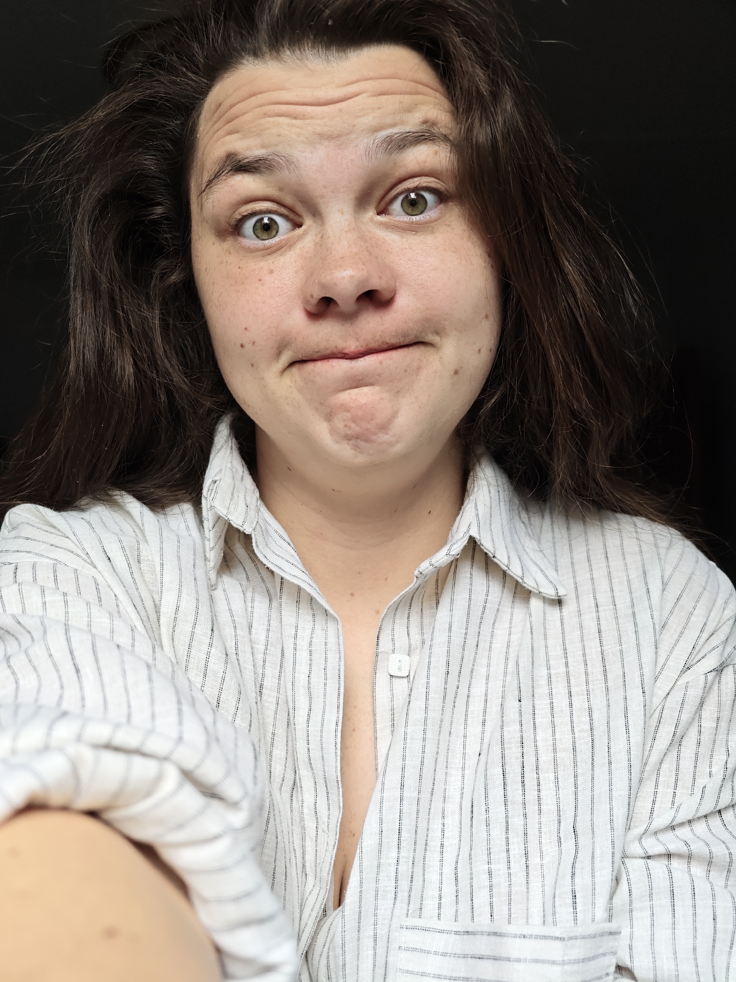 A medium close-up shot depicts a woman with fair skin, freckles, and light brown hair. She is wearing a white, vertically striped shirt with the top button undone. Her facial expression is quizzical, with raised eyebrows and a pursed mouth. The background is dark and indistinct. There is no indication of the location being Chaikovsky, Russia, or any other specific location.