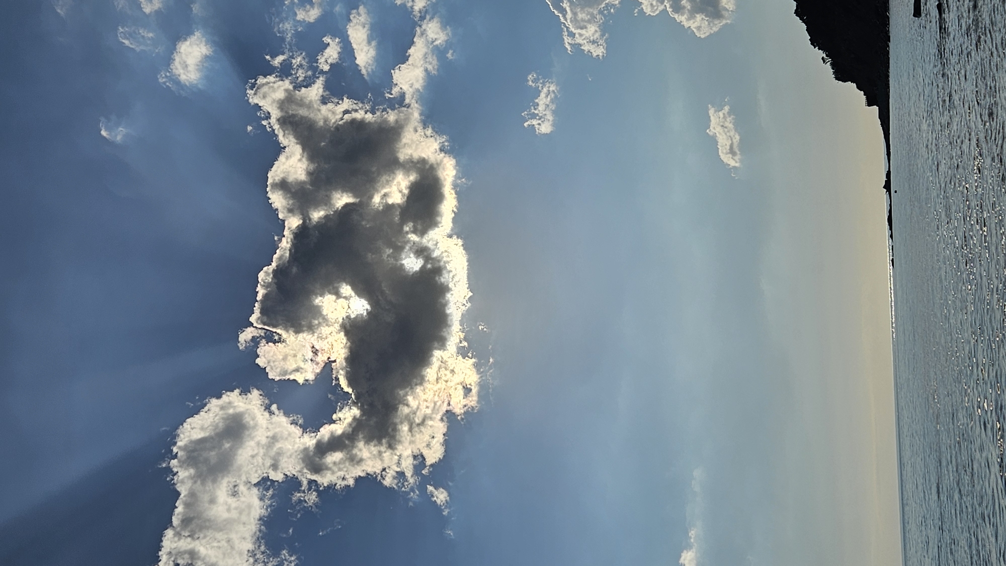 A wide-angle shot captures a dynamic sky over the calm waters of the Black Sea near Dyurso, Russia. The upper portion of the frame is dominated by a deep blue sky, transitioning to a lighter hue near the horizon. Sunlight pierces through a dramatic cloud formation, casting distinct rays downwards, creating a "god ray" effect. The cloud itself is a mix of dark, heavy masses and brightly illuminated edges, suggesting a powerful contrast of light and shadow.
Below the sky, the vast expanse of the sea stretches towards the horizon, its surface reflecting the sky's light with a shimmering effect. In the lower right corner, a silhouetted headland juts out into the sea, its dark form contrasting sharply with the bright water and sky. The headland appears to be covered in dense vegetation, with a small structure or pathway visible near its edge. The overall impression is one of natural beauty and the awe-inspiring power of the sky. There are no people or animals visible in the frame.