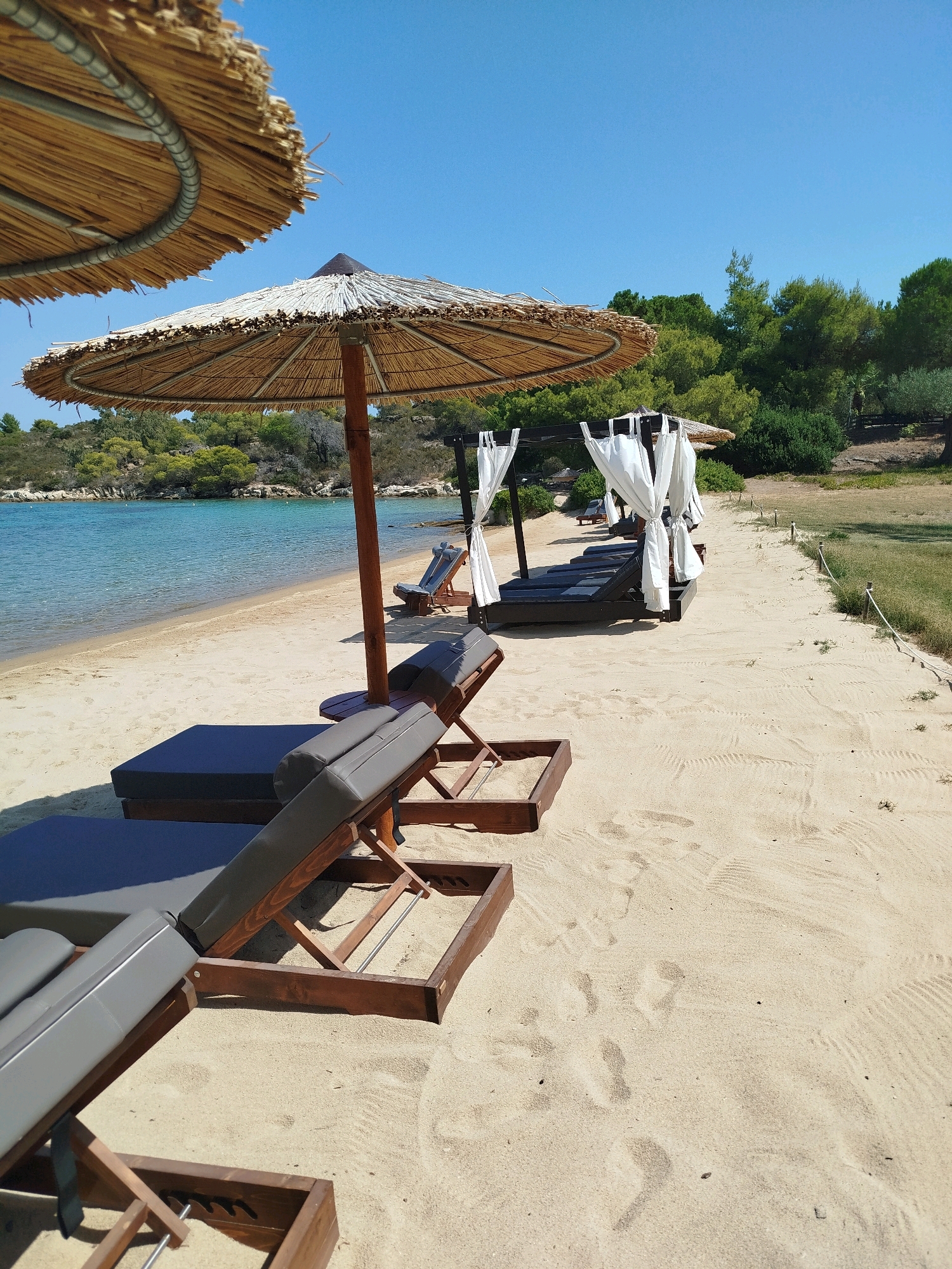This is a ground-level, eye-level shot of a beach on a sunny day in Sithonia Municipal Unit, Greece. The sand is light beige with some footprints and rake marks. In the foreground, there are two dark gray cushioned lounge chairs with wooden frames, positioned at an angle. Behind these, there is a large, dark-colored structure with white curtains draped around it, appearing to be a canopy or cabana, with more dark lounge chairs underneath. A straw umbrella with a wooden pole is situated near the lounge chairs, casting a shadow. Further back, the calm, turquoise water of the sea meets the sandy shore. Lush green trees and bushes line the background, with a clear blue sky overhead. There are no people visible in the scene.