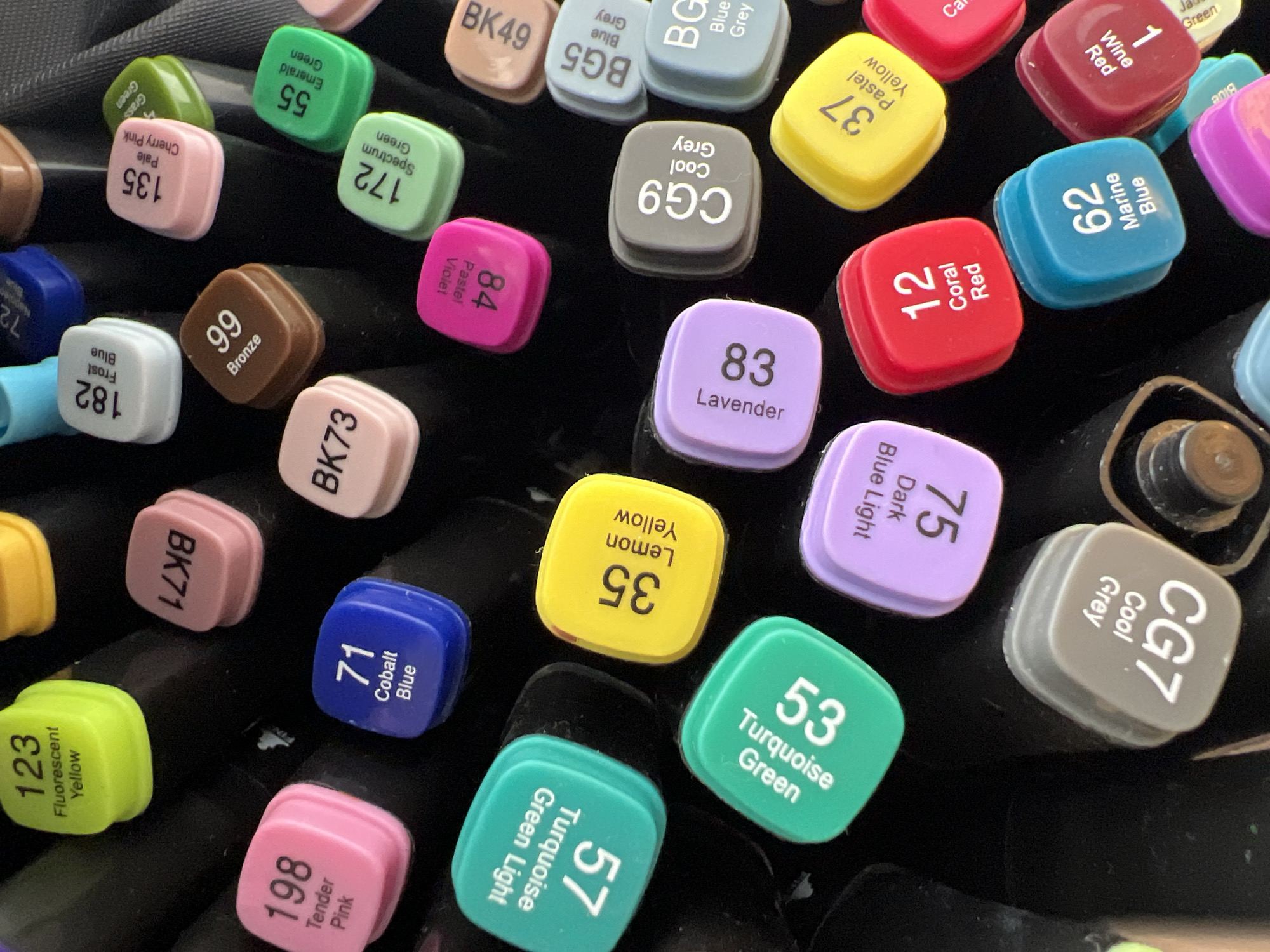 The image displays a close-up, overhead view of an assortment of marker caps, primarily in various shades of green, pink, yellow, blue, and grey. Each cap is square-shaped with rounded edges and features a label indicating a number and a color name. The caps are arranged haphazardly on a dark, textured surface, likely the barrel of the markers themselves. Some caps are more legible than others, with visible text such as "123 Fluorescent Yellow," "198 Tender Pink," "71 Cobalt Blue," "35 Lemon Yellow," "57 Turquoise Green Light," "53 Turquoise Green," "75 Dark Blue Light," "83 Lavender," "CG9 Cool Grey," "12 Coral Red," "62 Marine Blue," and "37 Pastel Yellow." The markers are likely artist-grade markers given the specific color names and numbering system, suggesting a creative or artistic context. The lighting appears to be artificial, casting subtle shadows on the surface. The image does not contain any discernible elements that would indicate a location in Minsk, Belarus, or any other specific geographical region. It is a still life composition focused on the details of the marker caps.