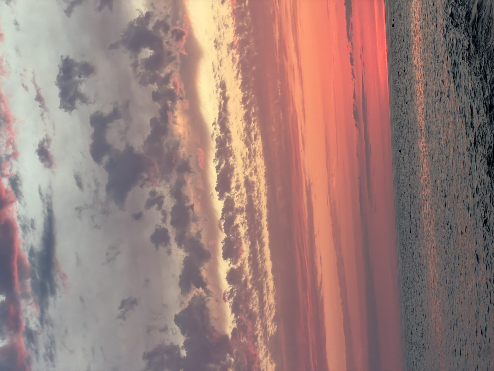 The image depicts a sunset over a body of water. The sky is filled with a gradient of colors, ranging from deep pinks and oranges near the horizon to lighter purples and grays higher up. Clouds are scattered across the sky, some appearing wispy and others more dense, catching the light and adding texture to the scene. The water below is dark and reflects the colors of the sky, with gentle ripples suggesting a calm surface. Several buoys or markers are visible in the water, breaking the expanse of the sea. The overall impression is one of a serene and picturesque natural landscape.