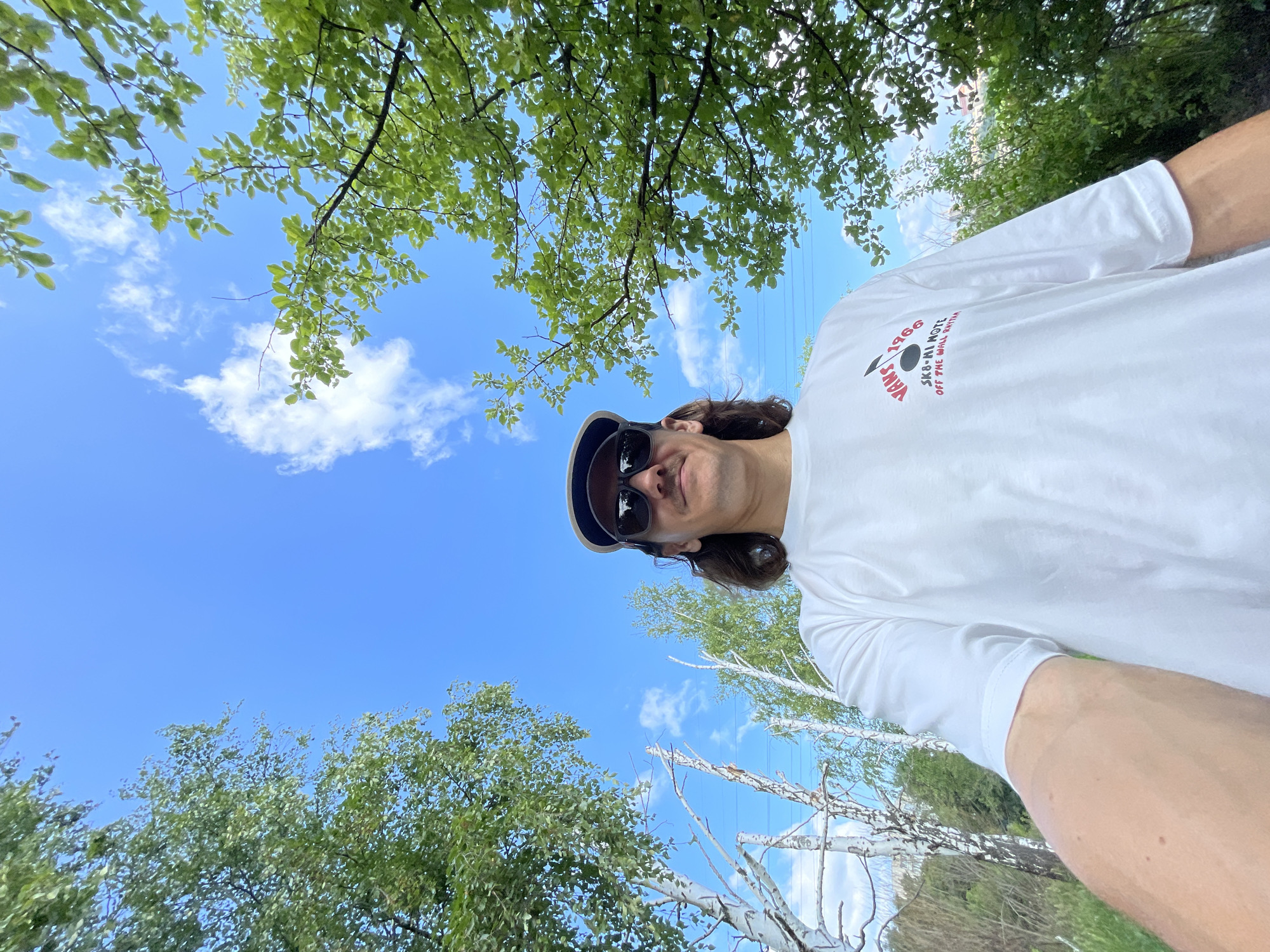 A low-angle, wide shot captures a man taking a selfie outdoors under a bright blue sky with scattered white clouds. The man is wearing a white t-shirt with a red and black graphic, sunglasses, and a baseball cap. He has shoulder-length brown hair and a mustache. Behind him, lush green trees frame the scene, with the upper branches of a deciduous tree in the foreground on the right. In the background, thin white birch trees are visible, along with power lines stretching across the sky. The overall setting suggests a natural, possibly park-like environment. The image was taken in Izhevsk, Russia.