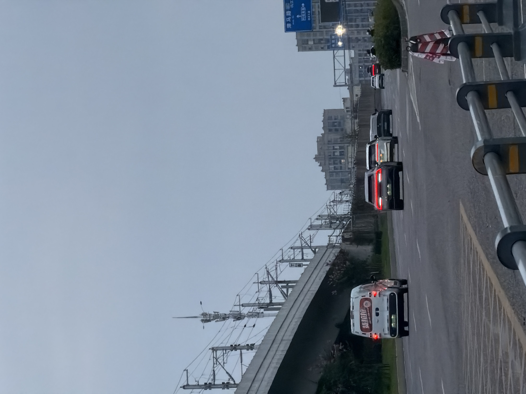 The image shows a street scene in Jiangjin, China, on an overcast day. In the foreground, a highway overpass with overhead power lines runs diagonally across the frame from left to right. Below the overpass, on the right side, there is a traffic sign with Chinese characters. A white van with red lettering is driving on the road in the left lane, facing away from the camera. Several other cars are also on the road, some with their brake lights illuminated. The road has lane markings and what appears to be a yellow and black striped barrier in the lower right corner. In the background, several multi-story buildings can be seen, and the sky is a uniform grey. The overall impression is that of urban infrastructure and transportation.