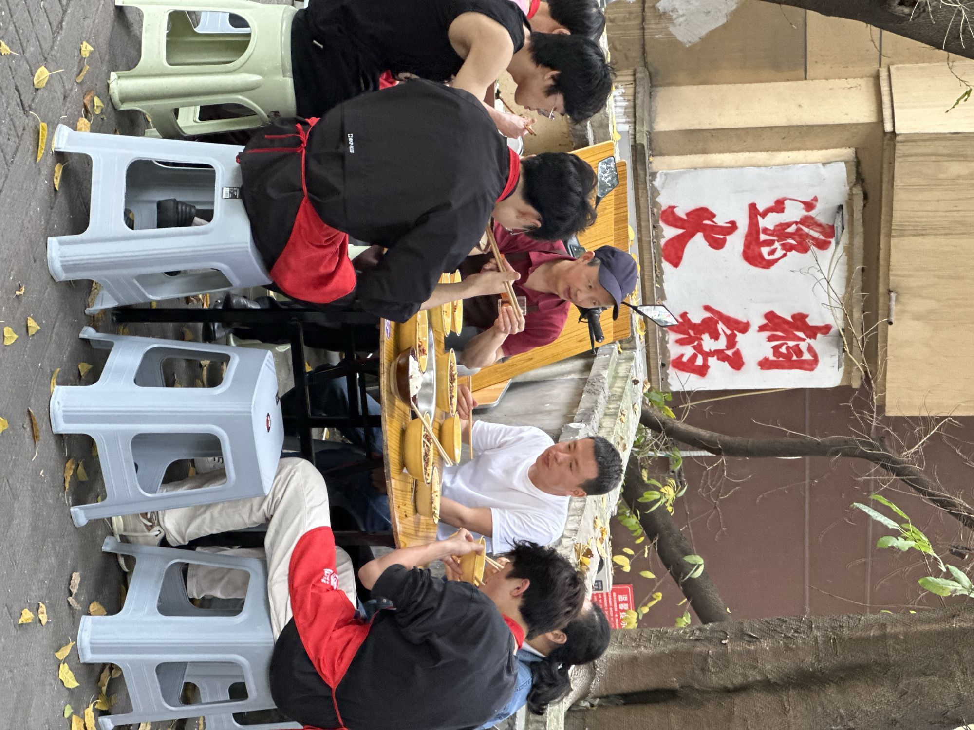 This image depicts an outdoor dining scene in Yuzhong District, China, featuring several individuals eating at a street-side establishment. At least seven people are visible, gathered around a long, narrow wooden table.
Most diners are seated on low plastic stools, varying in color from light grey to light green. Many of the younger men are wearing black shirts or jackets, some with red aprons, suggesting a possible uniform or group affiliation. Other individuals are dressed casually, including a man in a white t-shirt and another in a maroon polo shirt wearing a dark blue cap. All are actively eating from individual yellow bowls with chopsticks, indicating a communal meal. A small microphone or camera device is mounted on the table near the man in the cap.
The setting is an urban outdoor space with stone paving underfoot, adorned with scattered yellow leaves. In the background, there's a building wall, and tree branches with some green foliage are visible. A prominent white sign with bold red Chinese characters is affixed to the wall. The characters read "大邑" (Dàyì) at the top and "热卤" (Rèlǔ) below, which translates to "Hot Lu" or "Hot Braised Dishes," indicating the food specialty of the establishment. The scene takes place during the daytime under soft, possibly overcast, lighting conditions. The overall atmosphere is casual and focused on the shared dining experience.