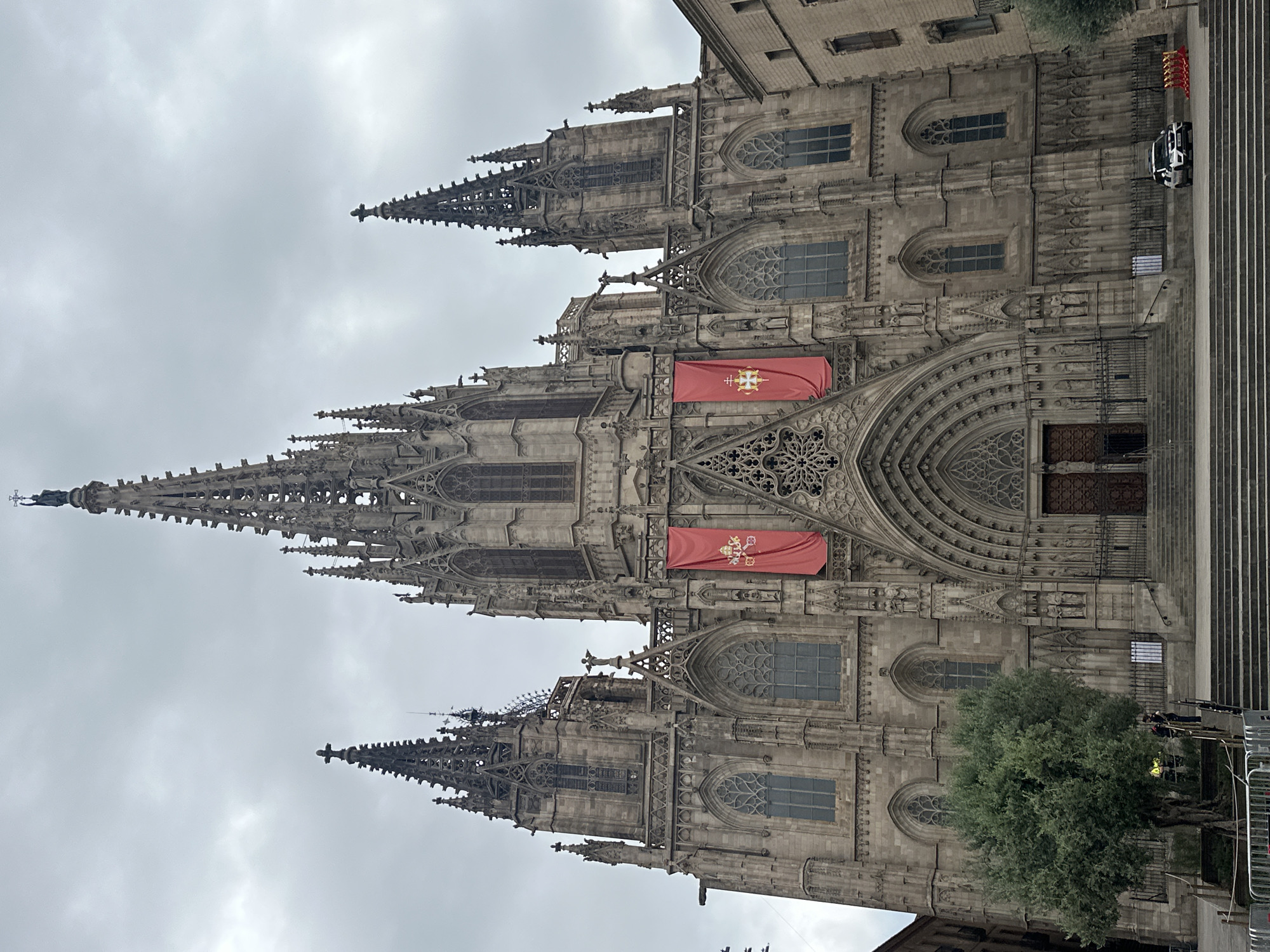 The image depicts the Barcelona Cathedral, a Gothic Roman Catholic cathedral located in Barcelona, Spain. The facade of the cathedral is made of stone and features intricate carvings, pointed arches, and spires reaching towards the overcast sky. Two large red banners with a golden crest are hung from the front of the building. In front of the cathedral, there are a set of wide stone stairs leading up to the entrance. To the left of the stairs, a mature olive tree stands behind a metal fence. A few people are visible near the base of the stairs, and a white car is parked to the right of the entrance. The scene appears to be in the late afternoon or early morning due to the muted lighting.