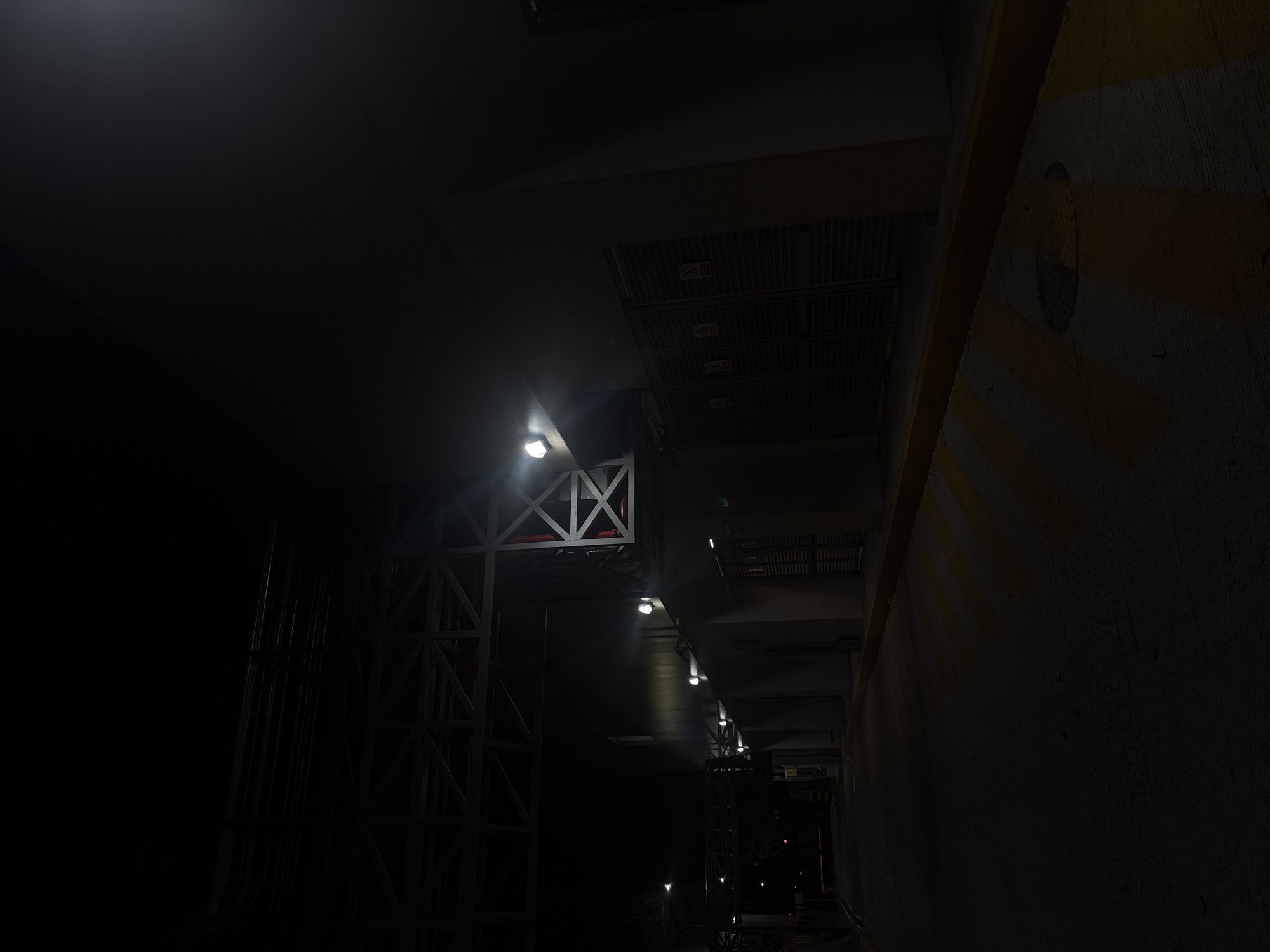 The image depicts a dimly lit industrial or infrastructure space. The scene includes a metal framework structure with vertical and diagonal supports, illuminated by a series of lights extending into the distance. Overhead are metal grilles and ductwork, possibly part of a ventilation or utility system. To the right is a concrete wall with yellow hazard stripes. The lighting is poor, obscuring details of the environment. There's no clear indication of people present or specific actions taking place. The environment seems to be enclosed, underground, or within a large structure. There is no direct evidence indicating a specific location in Puebla, Mexico.