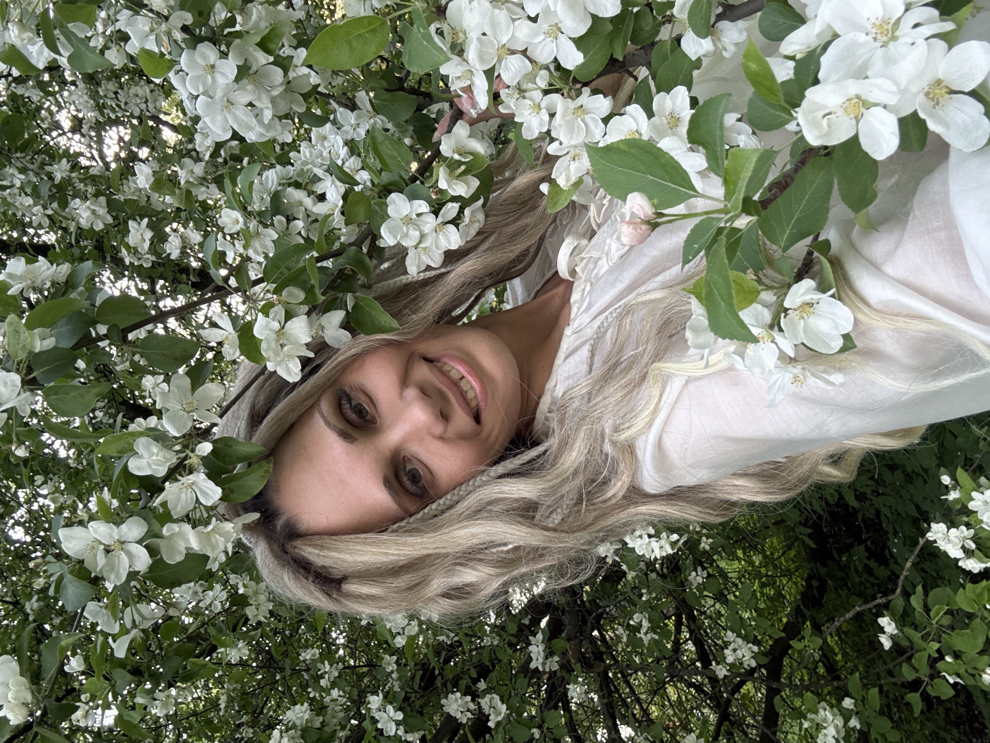 A woman is smiling amidst a blossom-filled tree, possibly an apple tree. She has long, wavy blonde hair and is wearing a light-colored top. The background is a mix of green leaves and more white blossoms. The location is given as the city of Шебунино, Russia.