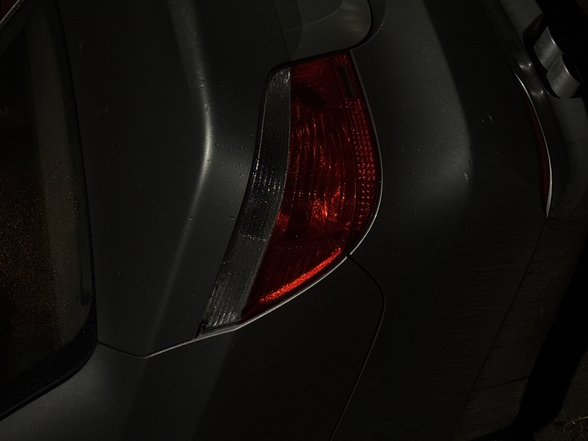 The image depicts the rear passenger side of a dark-colored car, possibly a hatchback, with its rear tail light visible. The tail light has a red and clear section. The car appears to be in a dimly lit environment, possibly at night. The presence of water droplets on the car's surface suggests recent rain or moisture. The scene's location within Varna, Bulgaria, is not evident from the image.