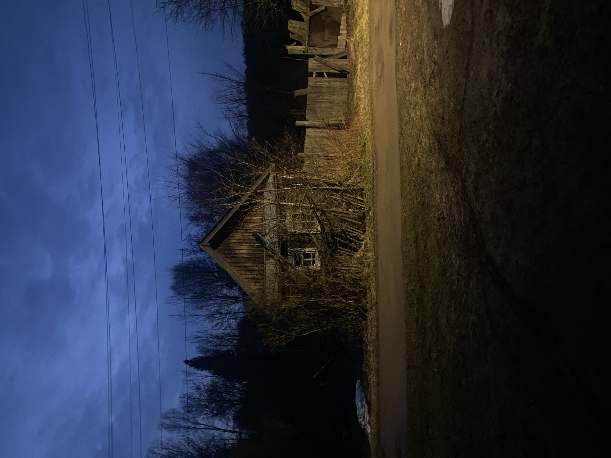 The image shows an abandoned, wooden house in what appears to be a rural setting. The house is located to the left of a dirt or gravel road. The time of day is dusk or night. The sky is a dark blue color. There are power lines visible above the house and the sky. The house is surrounded by bare trees and overgrown vegetation. There is a damaged wooden fence in the background. A dark, wooded area is also visible. The location is Zhigalki, Russia.
