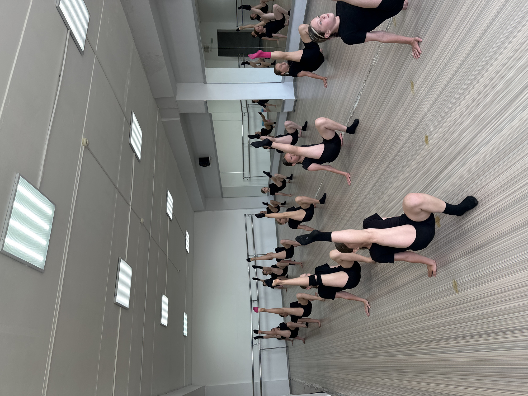 In a dance studio with light wood-paneled floors and a light grey wall, a group of young women are practicing dance. They are all dressed in black leotards and black shorts, with some wearing black socks and one wearing pink socks. The women are positioned in various yoga-like poses, with their bodies stretched and contorted. Some are on their backs with their legs in the air, while others are on their hands and knees with their backs arched. A large mirror runs along one wall of the studio, reflecting the dancers and the rest of the room. Above the dancers, ceiling lights are visible. The overall atmosphere appears to be one of focus and dedication, as the dancers are clearly engaged in a rigorous practice session. The lighting suggests it is daytime, and there are no visible weather cues. The location is a dance studio in Votkinsk, Russia.