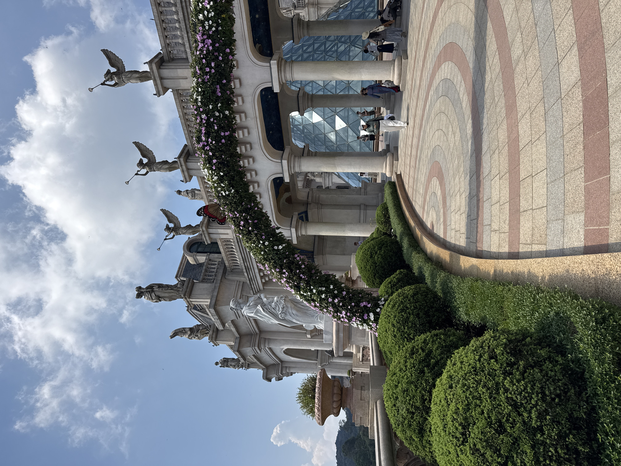 The image displays an exterior view of the French Village within Ba Na Hills SunWorld, situated in the Trường Sơn Mountains west of Da Nang city, Vietnam. The scene is illuminated by bright daylight under a partially cloudy blue sky.
The central element is a large, multi-story, European-style architectural complex constructed from light-colored stone or concrete. This structure features classical columns, arched passageways, balustrades, and a mansard roof. Numerous classical statues, including multiple winged figures holding trumpets positioned along the roofline, and draped figures, adorn the building. A prominent white statue of a draped female figure stands on a pedestal to the left of a main arched entrance.
A grand floral archway, adorned with white and light purple flowers, spans the main entrance, supported by large pillars. Through this archway, a significant multi-faceted glass pyramid structure is visible in the background. In the foreground, a curved paved walkway, constructed from interlocking rectangular paving stones in shades of light grey, reddish-brown, and off-white, leads towards the archway. This walkway is bordered by a manicured garden bed containing a row of neatly trimmed spherical green bushes and smaller ground cover. A large, terracotta-colored urn containing a green plant is present on a stone ledge to the far left.
Approximately 8-10 individuals are visible in the mid-ground and background, primarily within the archway area and near the glass pyramid. They are observed walking or standing, dressed in casual clothing suitable for a daytime public attraction. A large, artificial red and black butterfly decoration is affixed to the upper left section of the floral archway. Distant green forested mountains are visible on the horizon to the left.