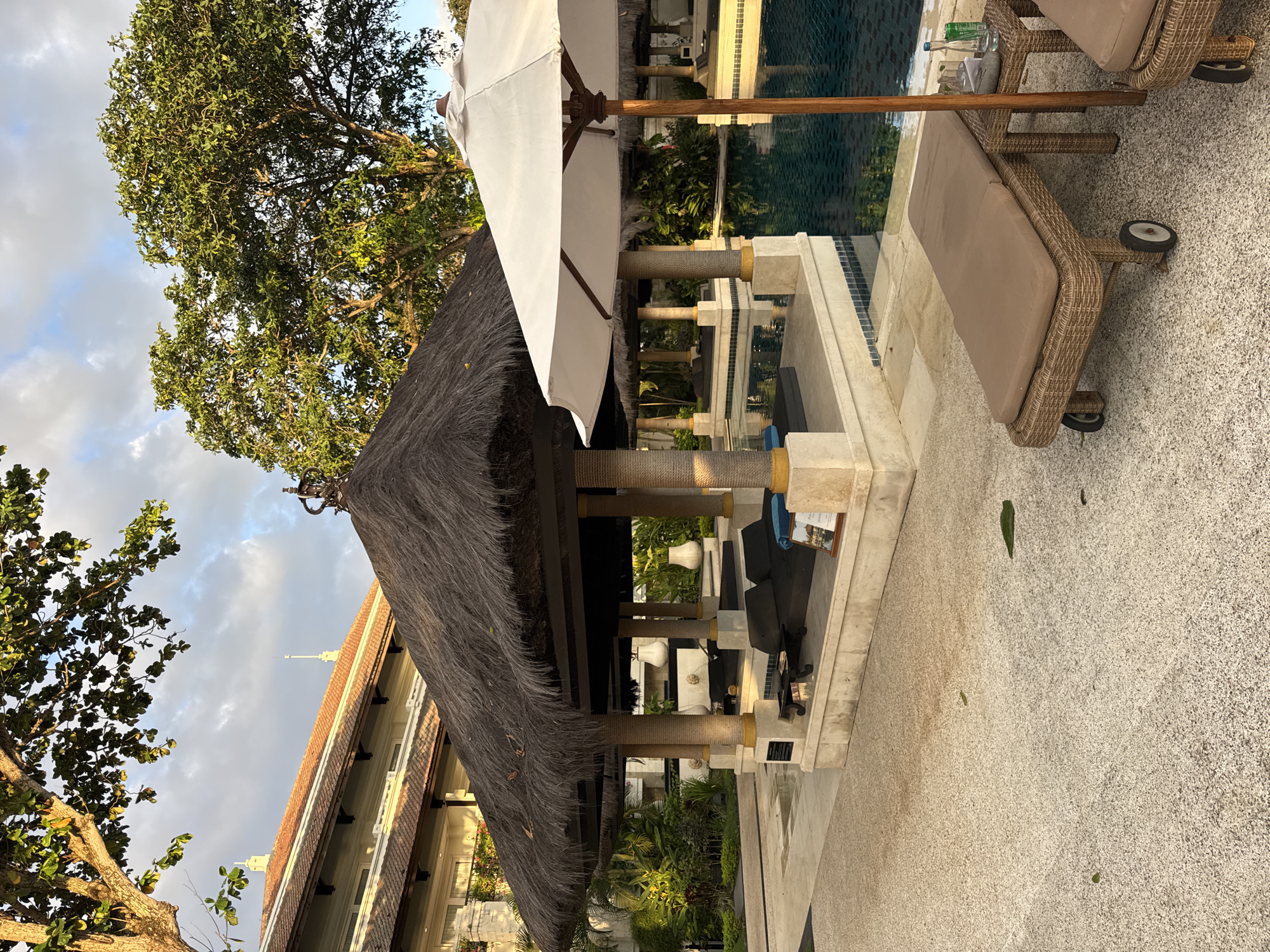 The image captures a serene outdoor setting in Kuta Selatan, Indonesia. A thatched-roof gazebo stands prominently, flanked by a white umbrella and a sun lounger made of wicker. The gazebo provides shade and shelter, while the umbrella offers additional sun protection. The lounger suggests a space for relaxation. A pool is visible in the background, with sunlight reflecting off its surface. Tropical vegetation surrounds the gazebo and pool area, enhancing the exotic ambiance. The ground is paved with small pebbles or stones. In the background, a building with a tiled roof can be seen, adding to the architectural context. The overall impression is one of a luxurious and peaceful resort environment. There are no people depicted in the image.