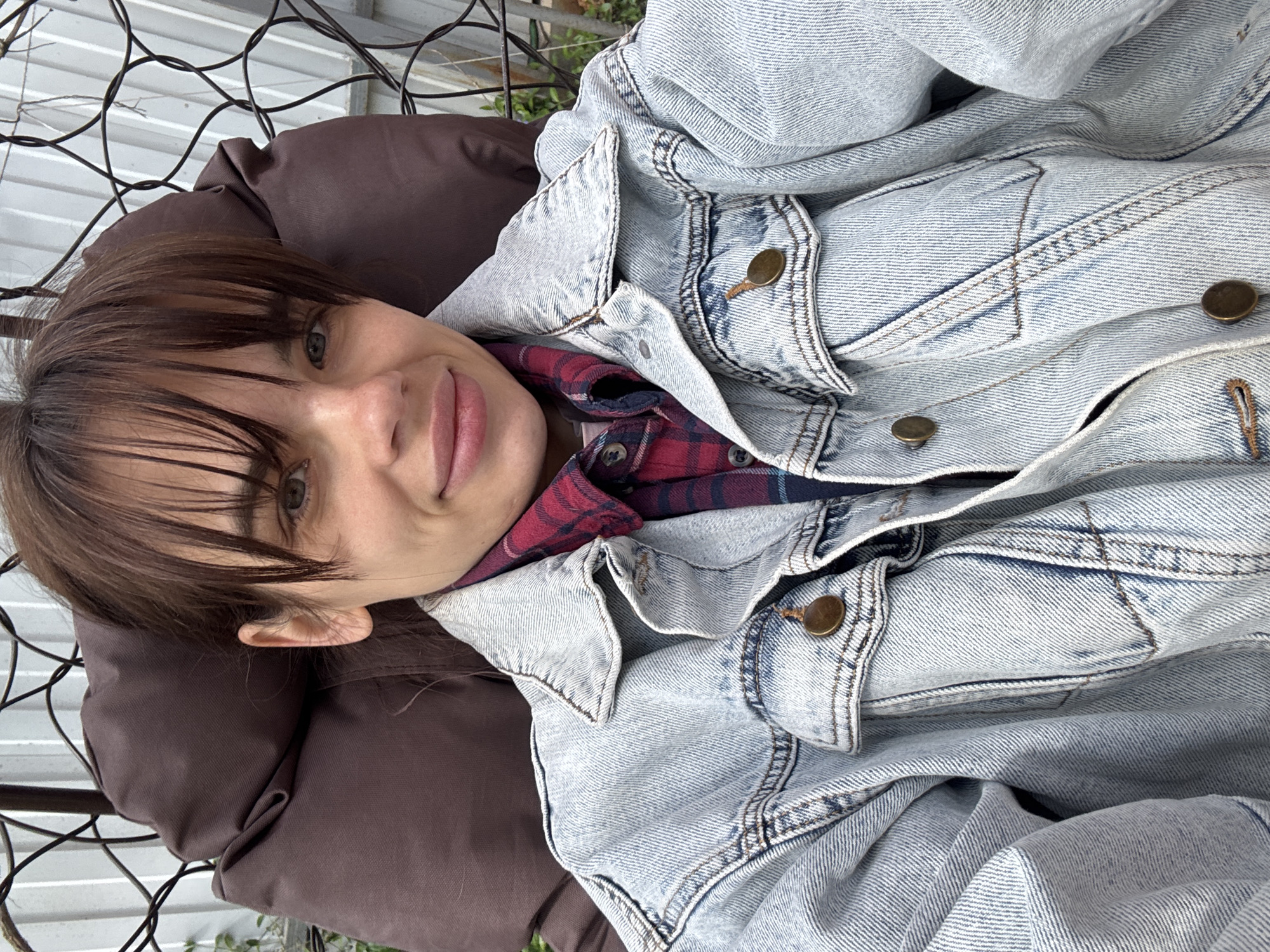 A woman with dark hair and bangs is lying in a brown cushion chair, wearing a blue denim jacket over a red and blue plaid shirt. The chair is part of a metal frame structure. The location is unknown, but the prompt requested the city of Rostov-on-Don, Russia.