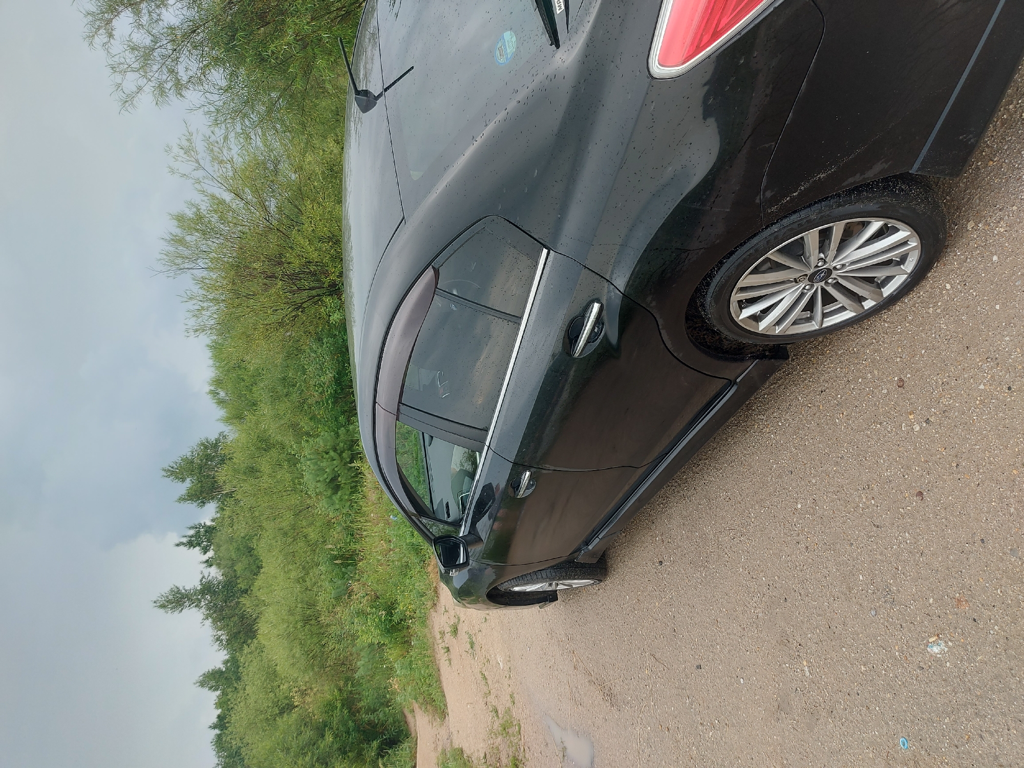 A black Subaru sedan is parked on a dirt road in Parunovka, Russia, surrounded by lush green foliage. The car is viewed from the rear passenger side, and its glossy black paint is speckled with raindrops. The road appears to be a path leading through a wooded area, with a small puddle of water visible on the left side of the frame. The sky is overcast, suggesting a cloudy or rainy day. The environment is natural and rural, with no visible buildings or other man-made structures in the immediate vicinity.