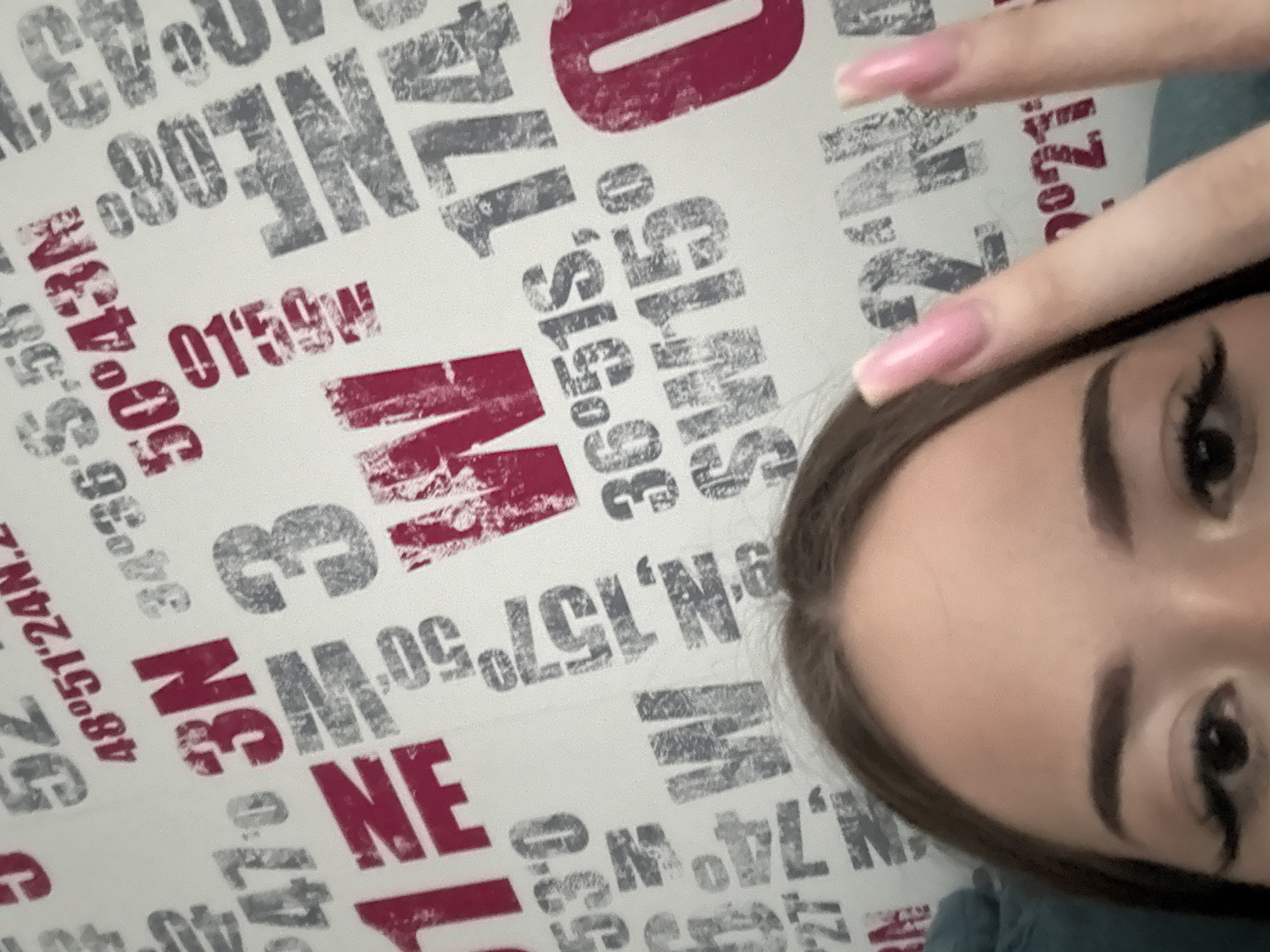 A close-up selfie shows a person's face with dark eyes, eyebrows, and brown hair. The person is holding up two fingers with pink manicured nails. The background features a pattern of latitude and longitude coordinates in red and gray lettering on a white surface.